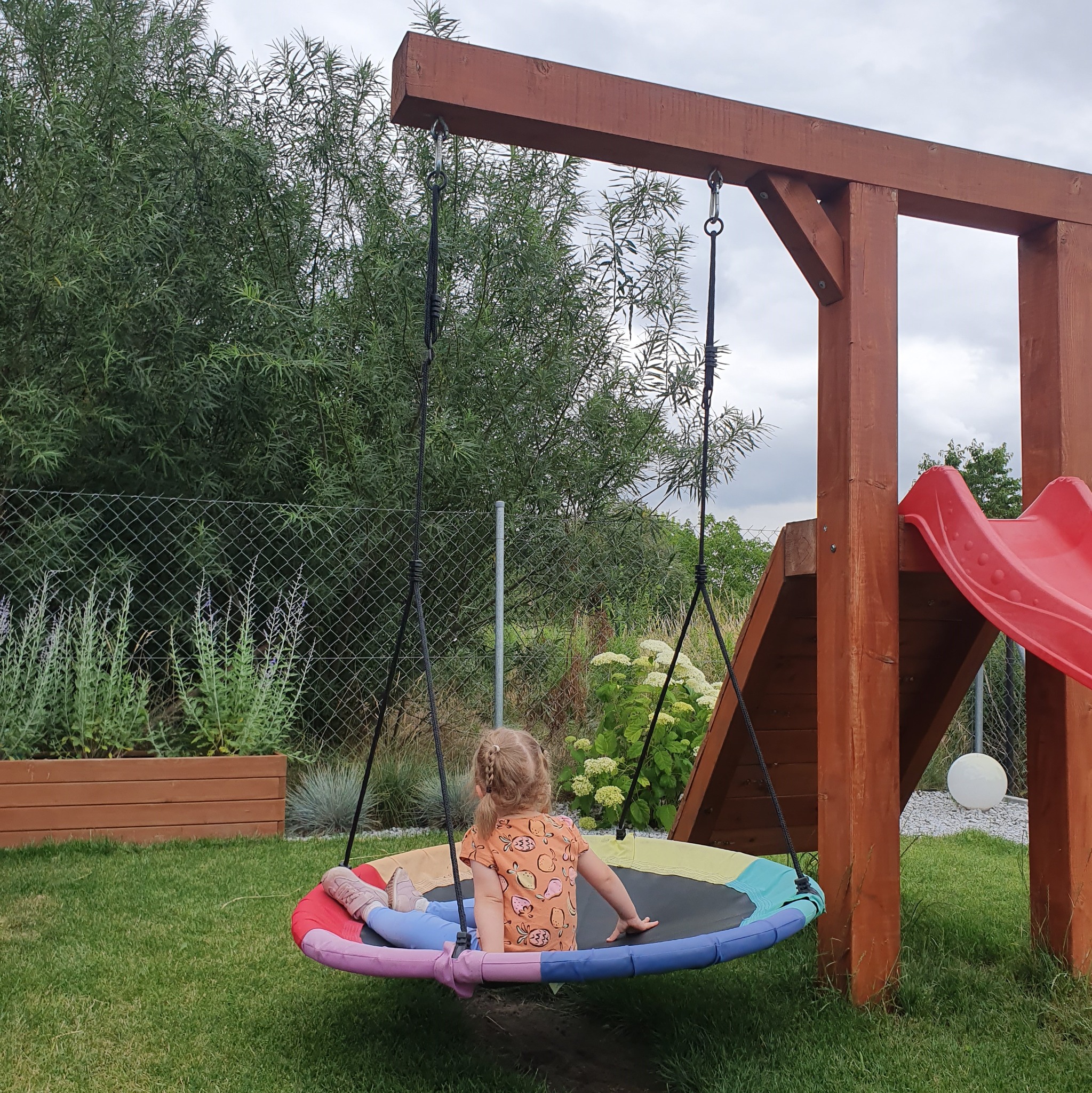 Dziewczynka siedzi na okrągłej, kolorowej huśtawce zawieszonej na drewnianej konstrukcji placu zabaw z czerwoną zjeżdżalnią, widok z tyłu, na trawniku, w tle zielone krzewy i ogrodzenie z siatki.