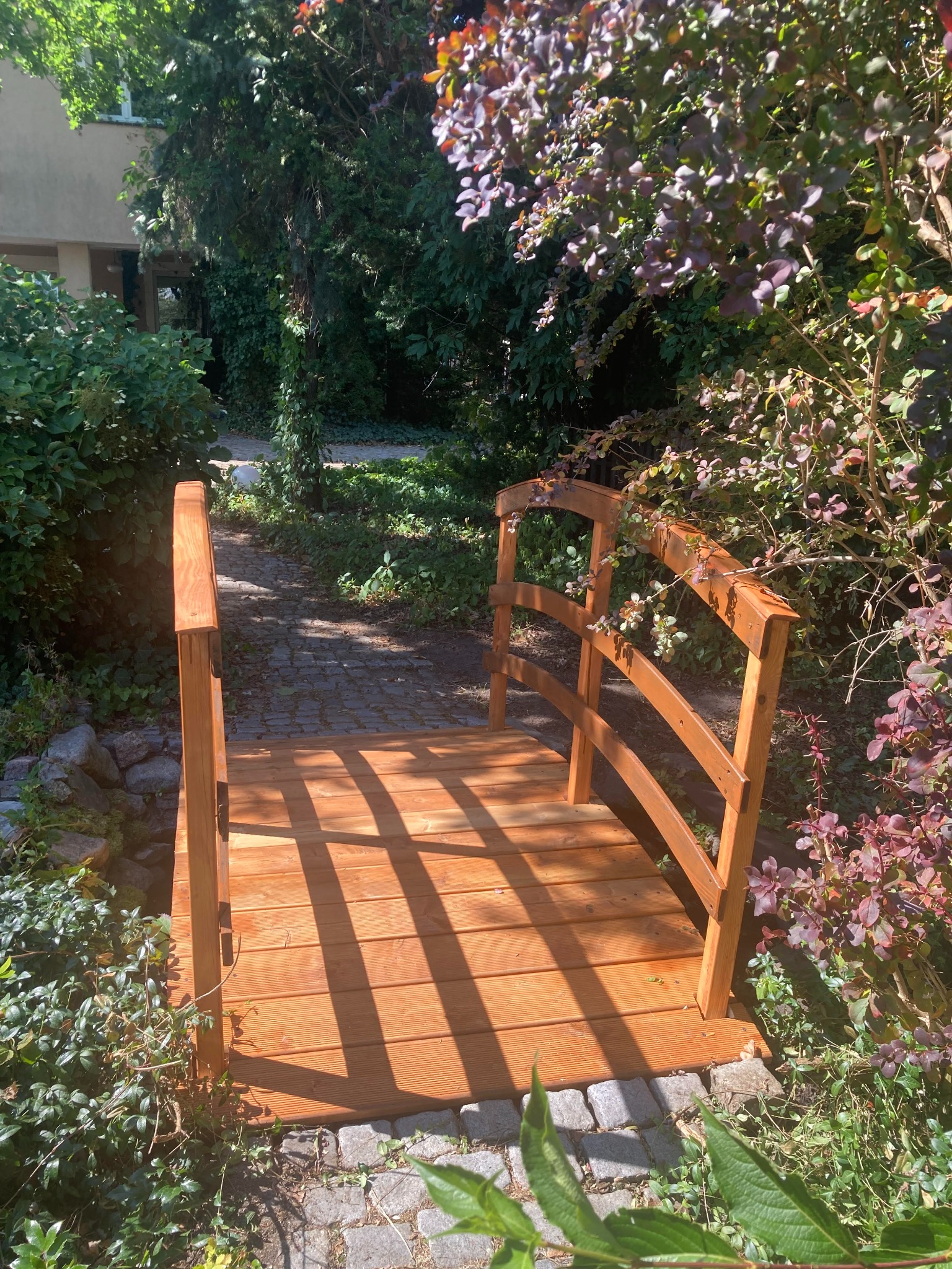 Mały drewniany mostek nad ścieżką w ogrodzie, z widoczną strukturą drewna i cieniem rzucanym przez barierki, otoczony zielenią i kamieniami.