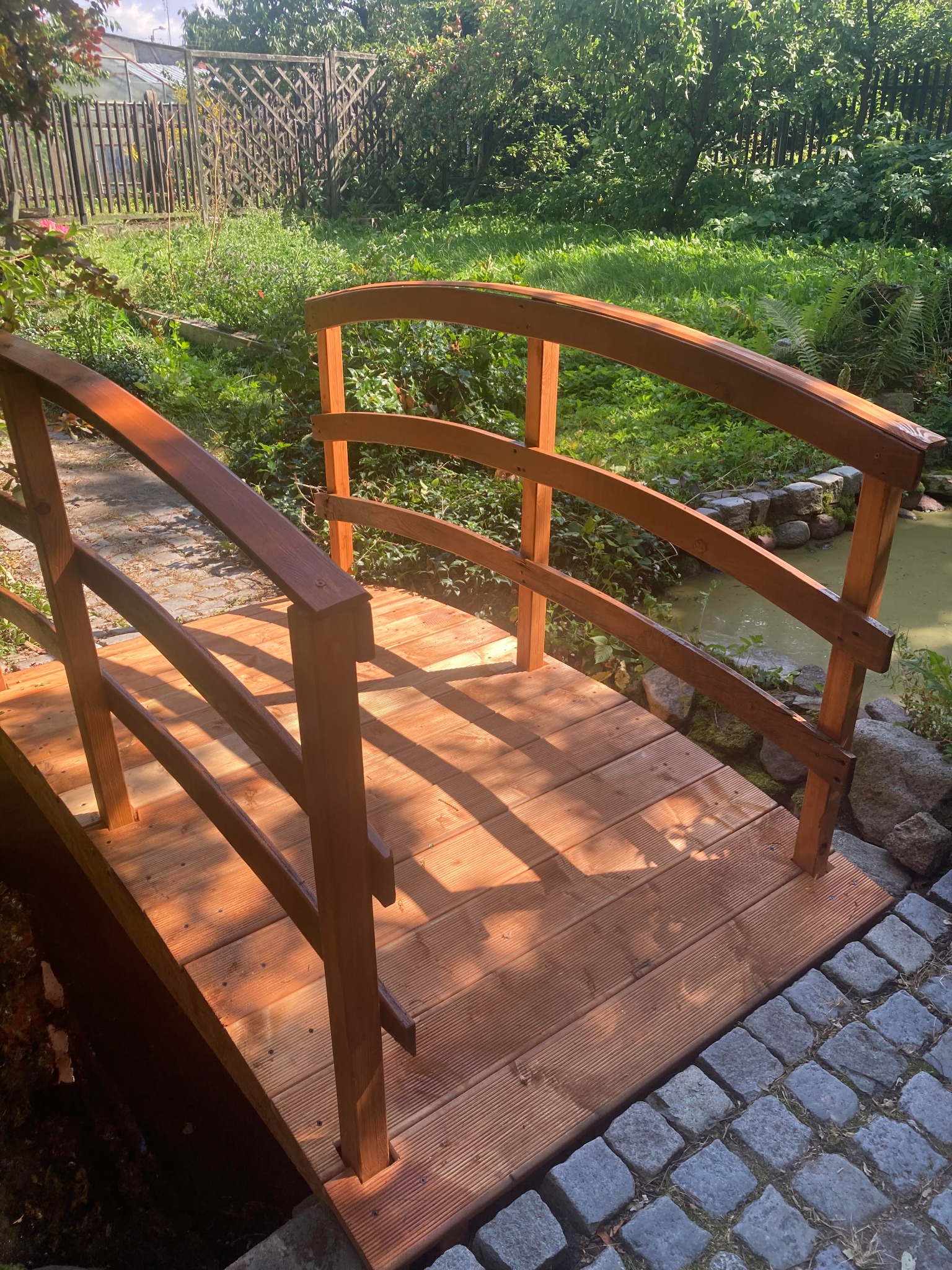 Mały drewniany mostek z barierkami prowadzący nad stawem w ogrodzie, wykonany z desek ryflowanych, obok ścieżka z kostki brukowej.