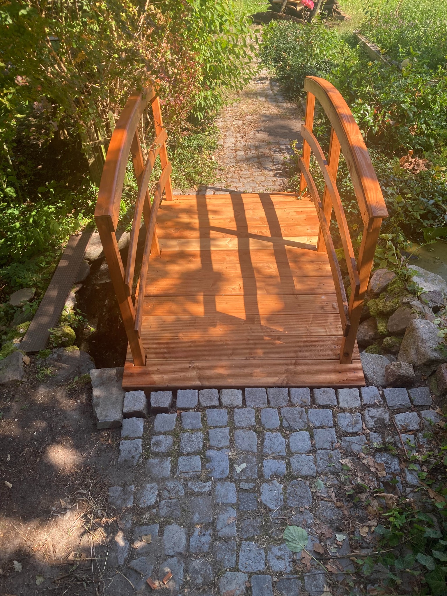 Mały drewniany mostek z balustradami, prowadzący przez strumyk w ogrodzie, z kamienną ścieżką po obu stronach i bujną roślinnością w tle, oświetlony popołudniowym słońcem.