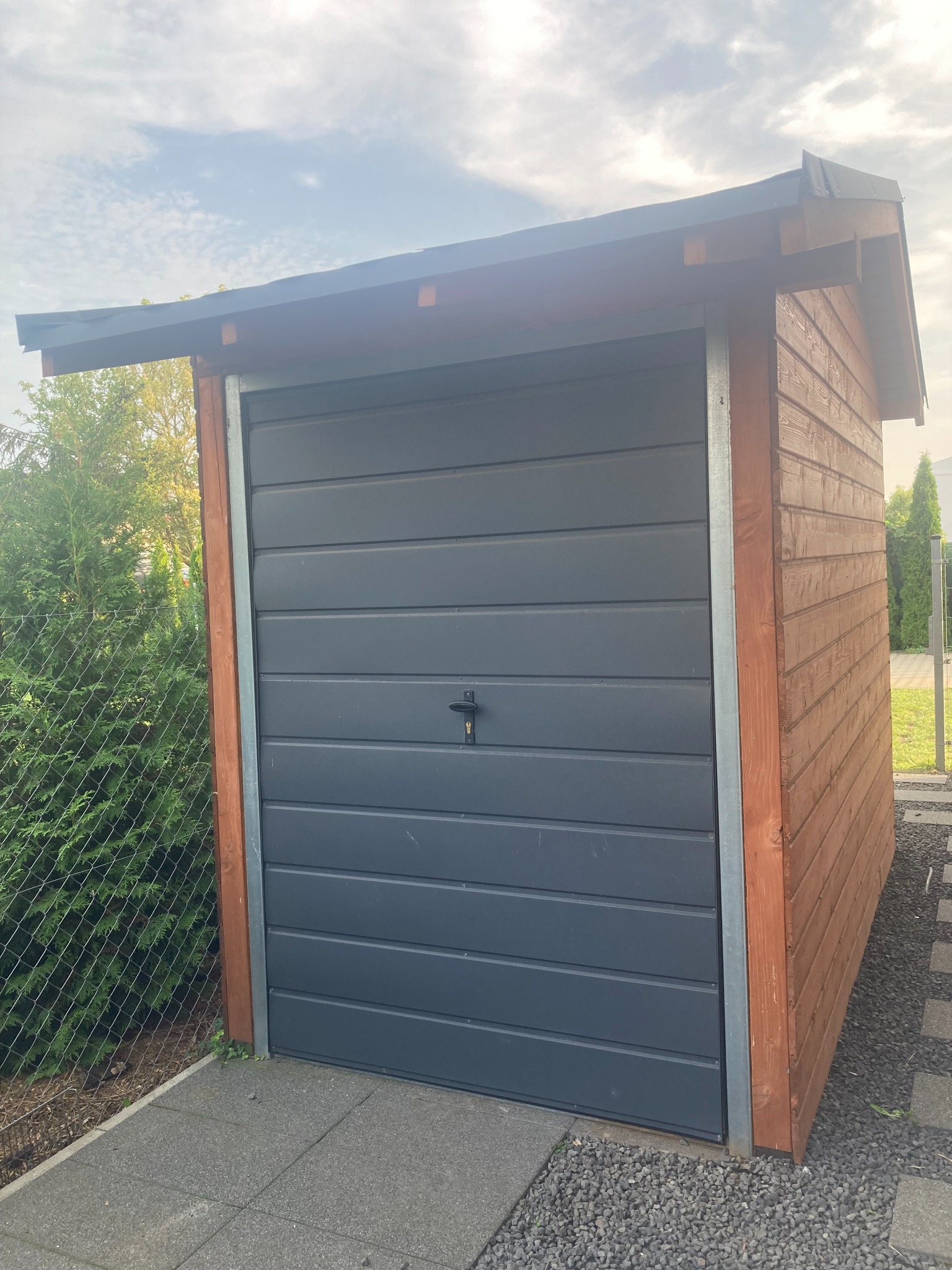 Mała, drewniana szopa z szarą, segmentową bramą garażową i płaskim dachem, otoczona kostką brukową i żwirem, widoczna z zewnątrz w ogrodzie.