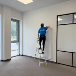 Pracownica na drabinie czyści biały grzejnik w jasnym biurze. Widoczne okna, wykładzina i przeszklona ściana. Ubrana w niebieską koszulkę firmową.