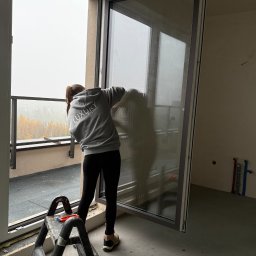 LUXURY EXPERT SERVICE DARIA LESZKO - Osoba w szarej bluzie z napisem 'Luxury Expert' myje duże okno balkonowe w nowym budynku. Widoczna drabina, wiadro i narzędzia. Za oknem jesienny krajobraz.