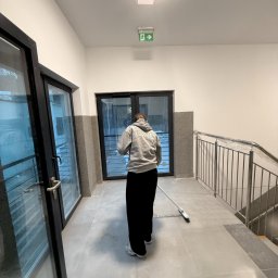 LUXURY EXPERT SERVICE DARIA LESZKO - Osoba w szarej bluzie z napisem 'Luxury' zamiata szarą posadzkę w korytarzu z oknami i metalową barierką schodową. Widoczny znak ewakuacyjny.