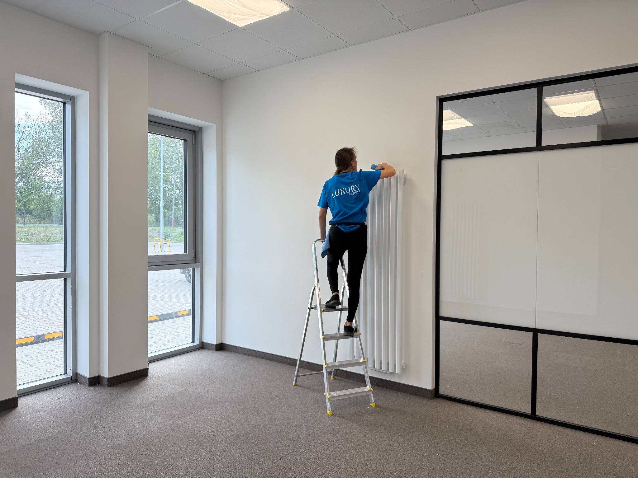 Pracownica na drabinie czyści biały grzejnik w jasnym biurze. Widoczne okna, wykładzina i przeszklona ściana. Ubrana w niebieską koszulkę firmową.
