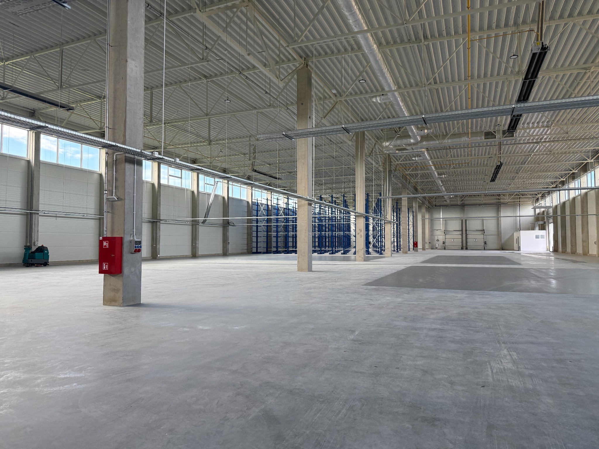 Wnętrze hali magazynowej po czyszczeniu posadzki, widoczny betonowy filar z czerwoną skrzynką i automat do mycia podłóg w tle. Metalowa konstrukcja dachu.
