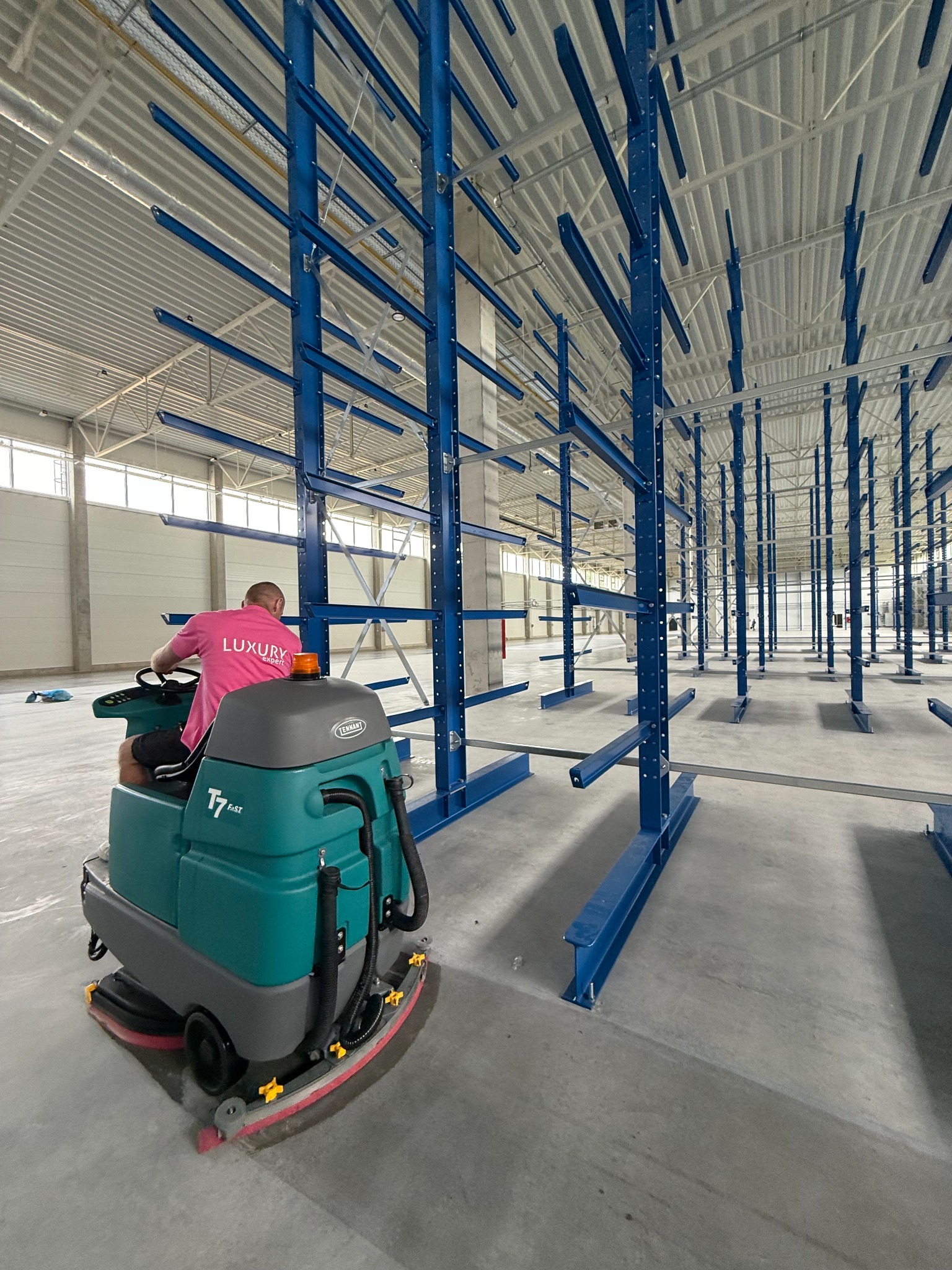 Operator na maszynie czyszczącej Tennant T7 FaST myje posadzkę w hali magazynowej z niebieskimi regałami. Widoczne ślady mycia.