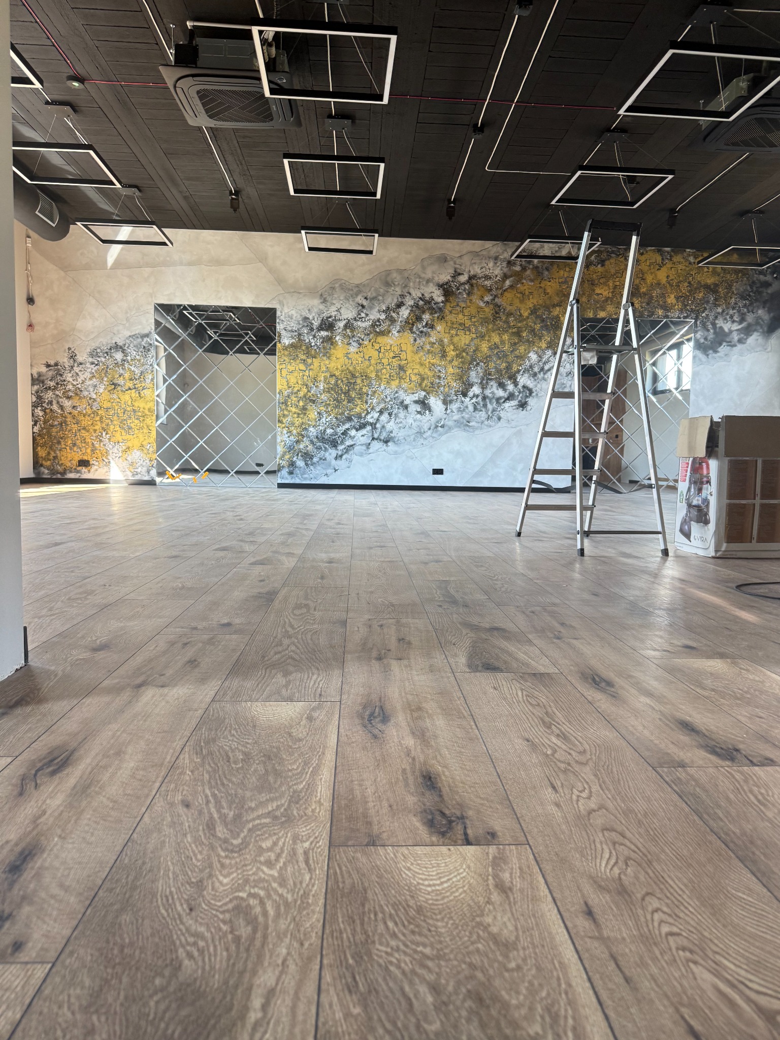 Wykończone wnętrze biura z drewnianą podłogą, dekoracyjną ścianą w odcieniach szarości i złota oraz lustrami. Widoczna drabina i karton wskazują na prace wykończeniowe.