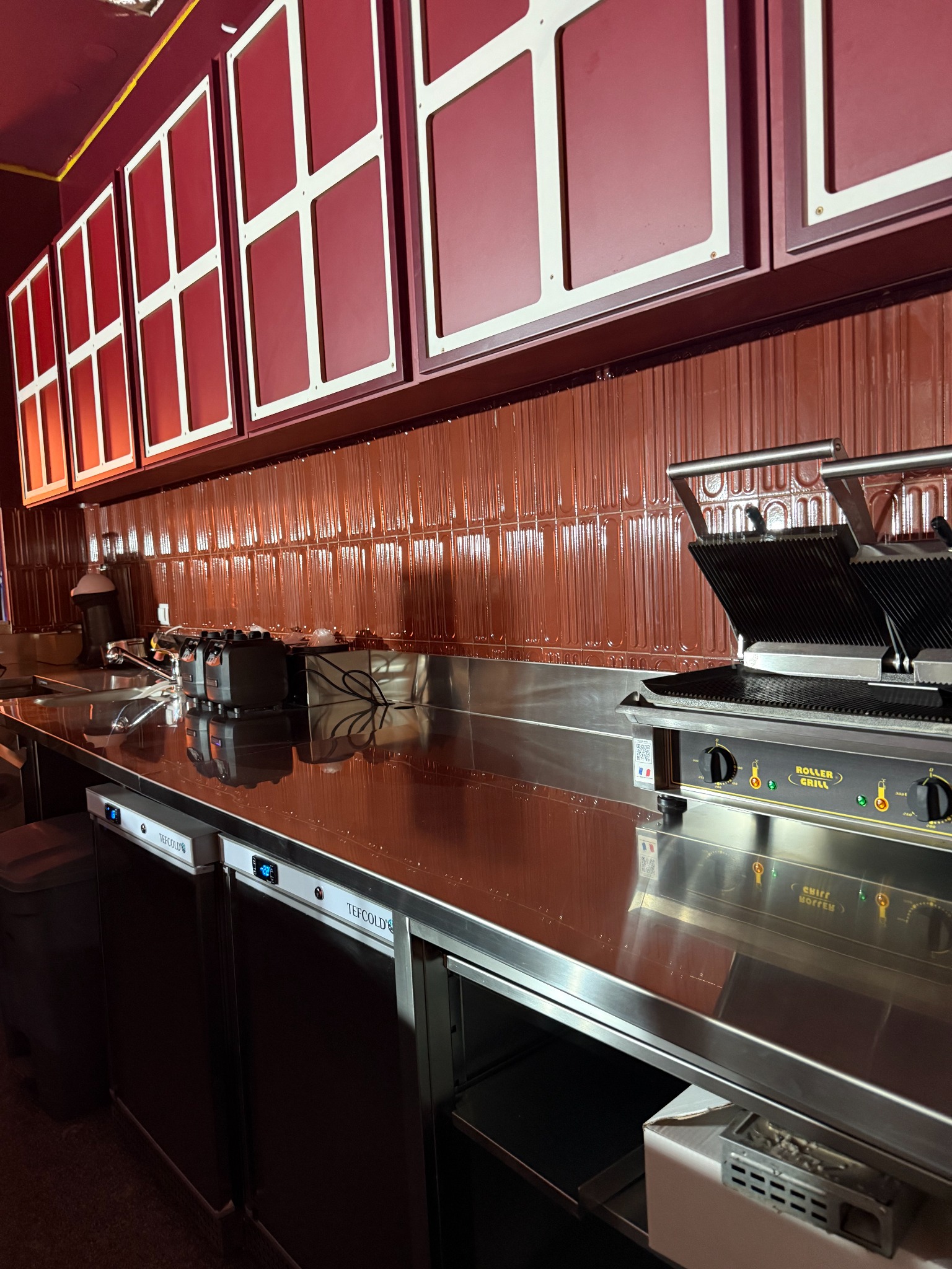 Profesjonalna kuchnia lokalu gastronomicznego z błyszczącym blatem, grillem i chłodziarkami TEFCOLD. Czerwone płytki i okna w tle. Widoczne detale wyposażenia.
