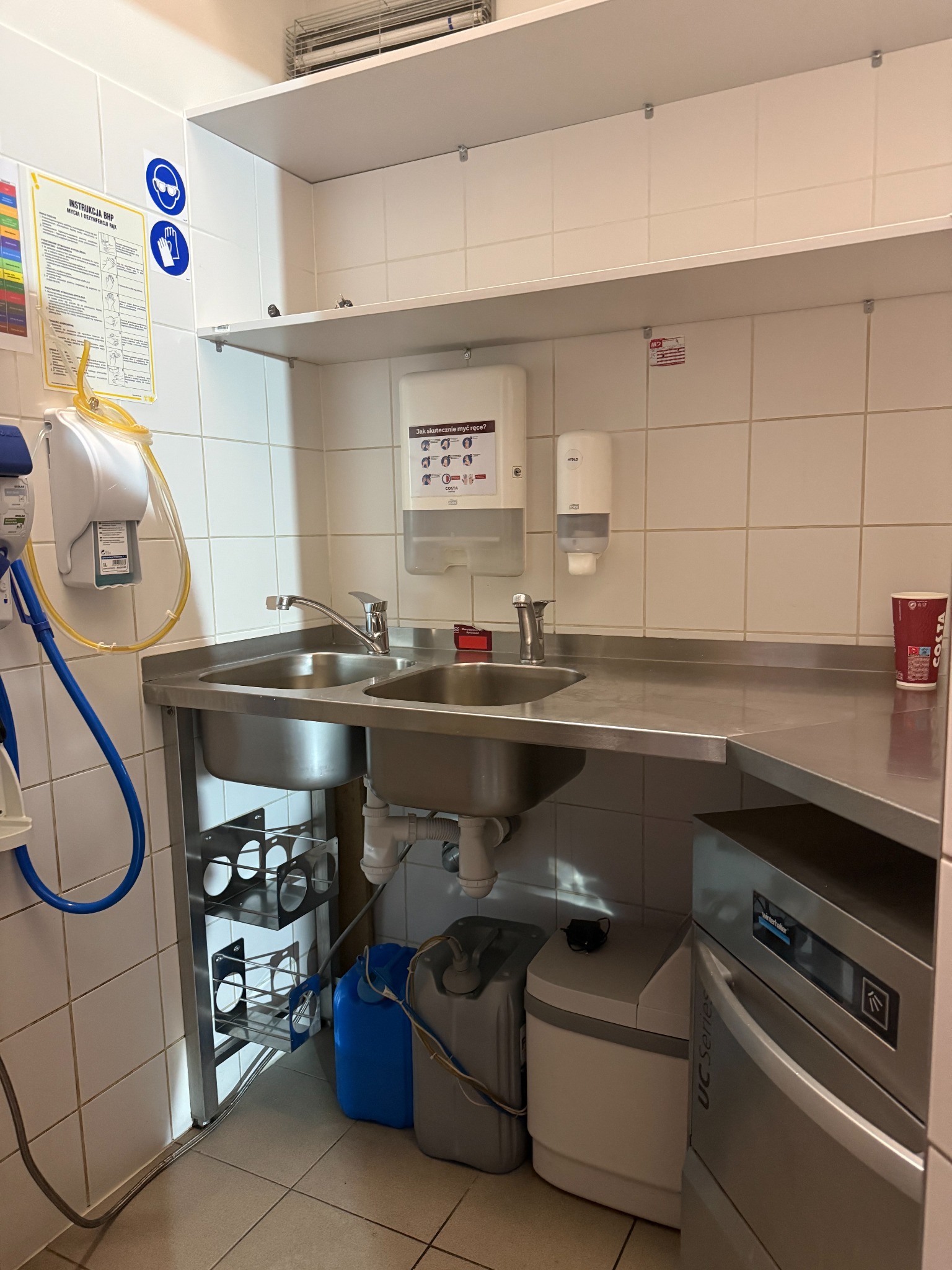 Profesjonalna kuchnia z nierdzewną umywalką, dozownikiem mydła, instrukcją BHP i zmywarką Winterhalter UC Series. Czyste, białe płytki i półki. Kubek Costa na blacie.
