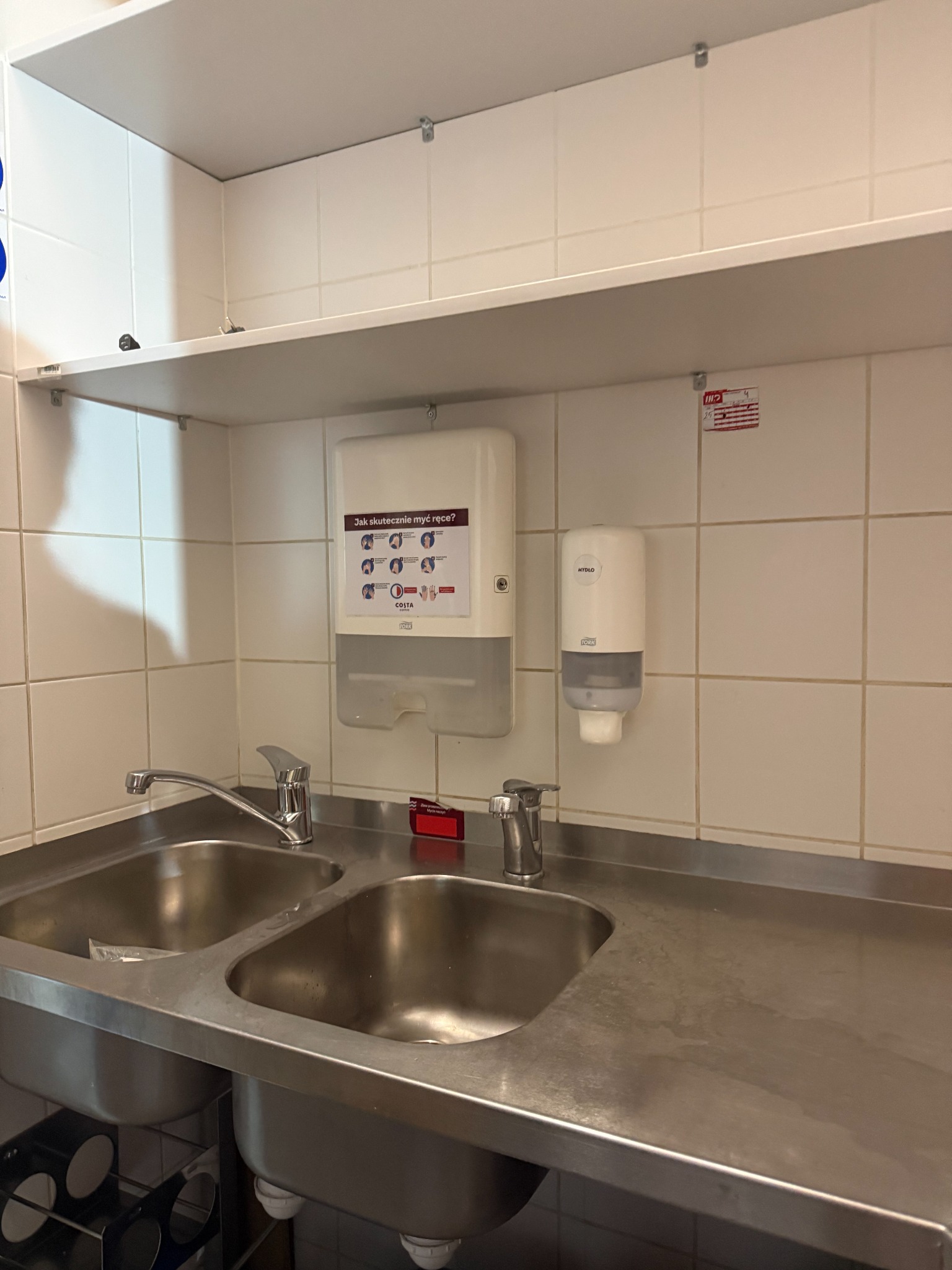 Stalowa umywalka dwukomorowa w lokalu gastronomicznym, dozownik mydła i instrukcja mycia rąk na tle białych płytek. Widoczne ślady użytkowania na powierzchni.
