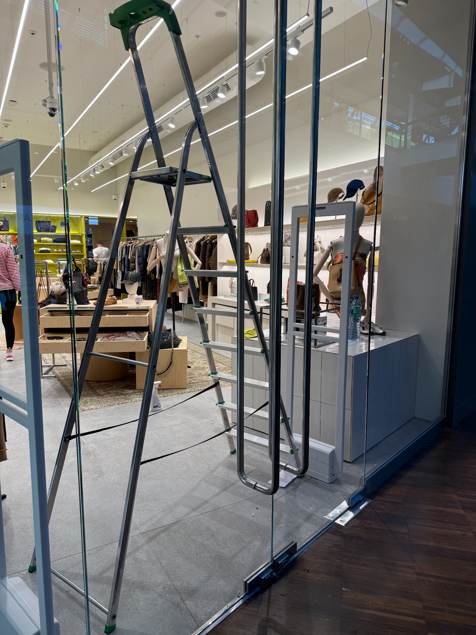 Składana drabina w sklepie odzieżowym, widoczna przez szklane drzwi. W tle manekiny i ubrania. Przygotowania do czyszczenia lub zmiany ekspozycji.