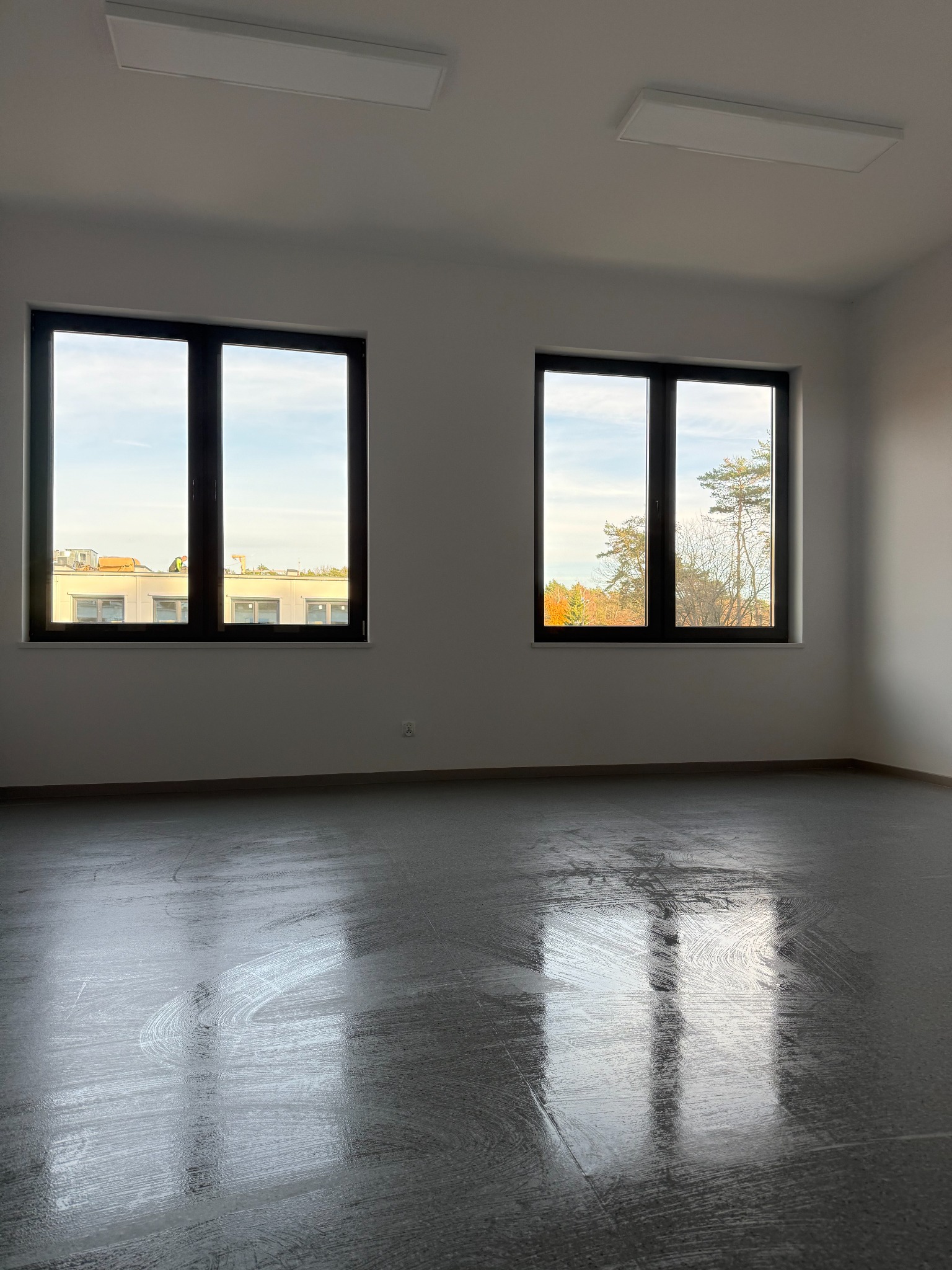 Wnętrze po sprzątaniu: jasne ściany, dwa okna z widokiem na zewnątrz, czysta podłoga z refleksami światła. Widoczne ślady po czyszczeniu podłogi.