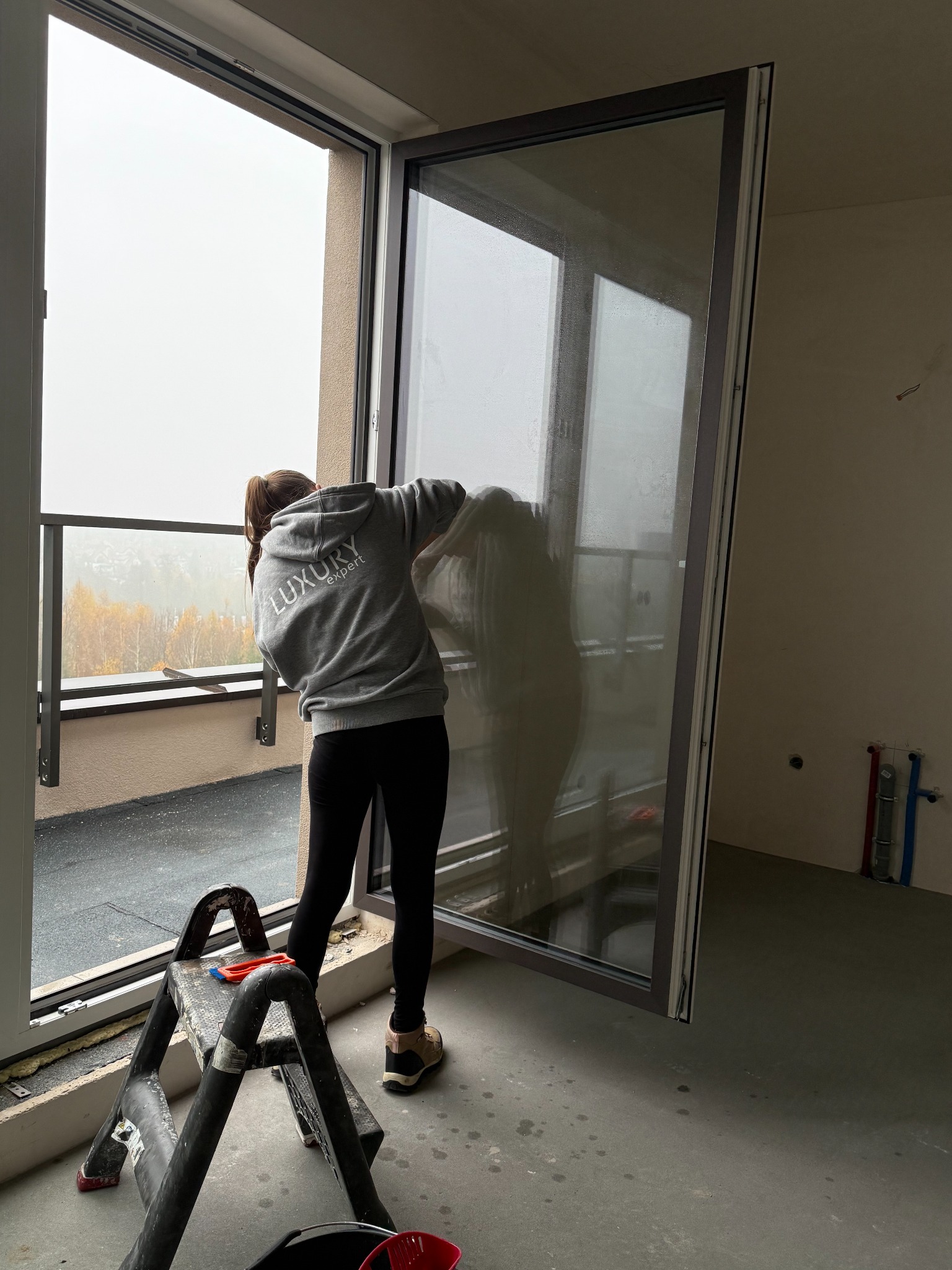 Osoba w szarej bluzie z napisem 'Luxury Expert' myje duże okno balkonowe w nowym budynku. Widoczna drabina, wiadro i narzędzia. Za oknem jesienny krajobraz.