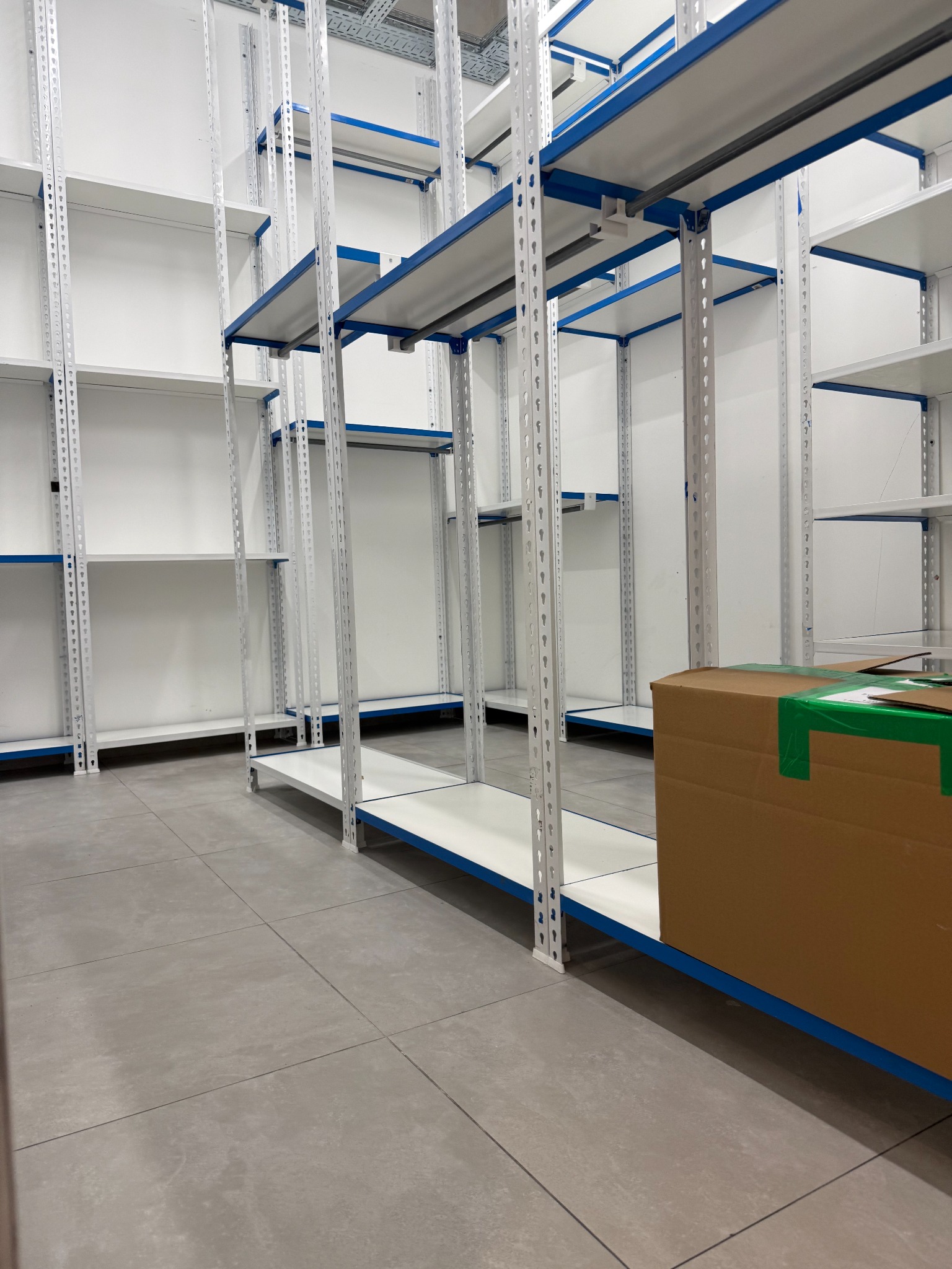 Półki magazynowe z niebieskimi akcentami, częściowo puste, z kartonem na pierwszym planie. Widok na czystą, szarą podłogę i białe ściany. Perspektywa z dołu.