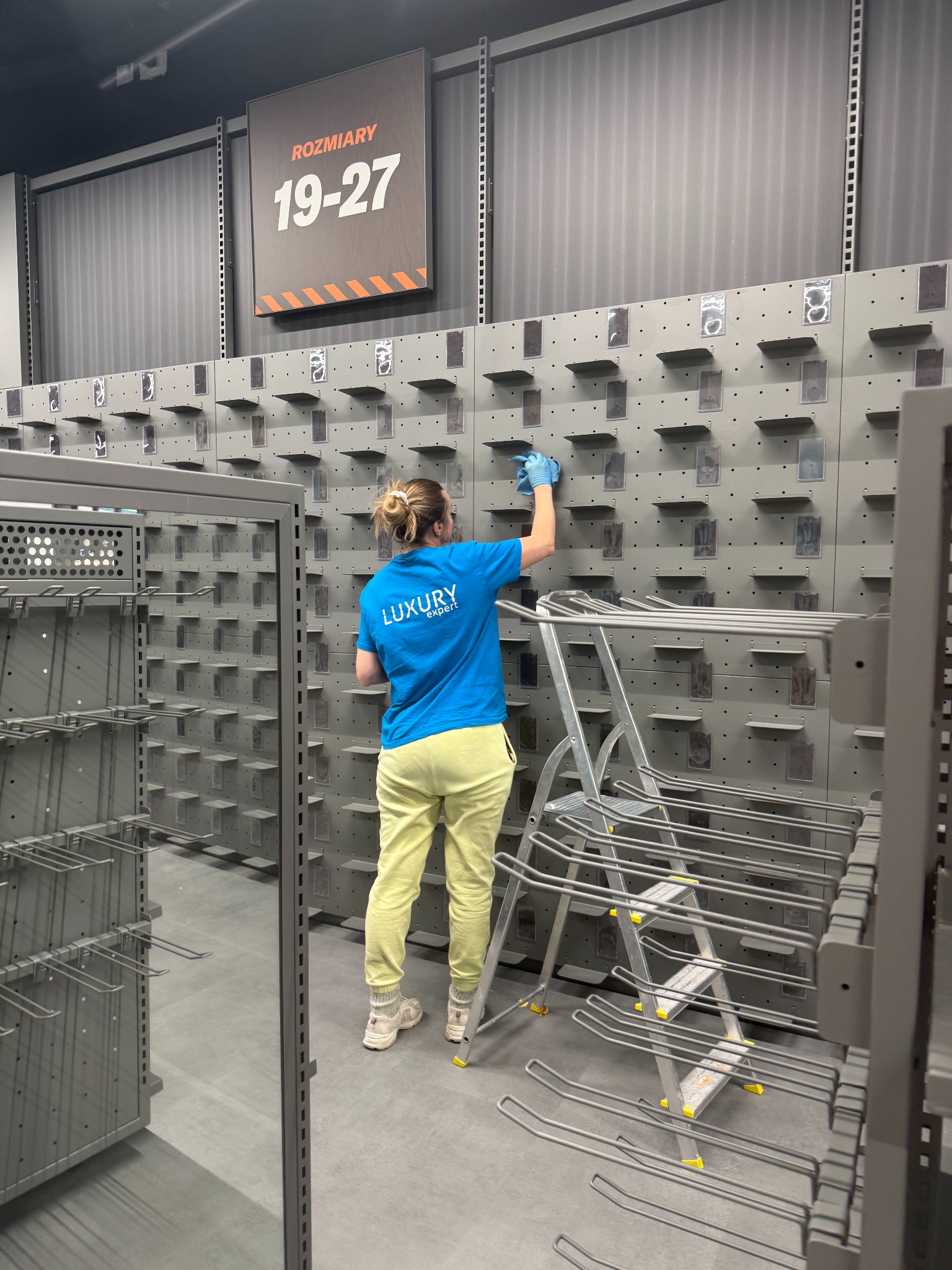 Pracownica w niebieskiej koszulce czyści półki w sklepie odzieżowym, używając drabiny. Widoczny napis 'Rozmiary 19-27'.