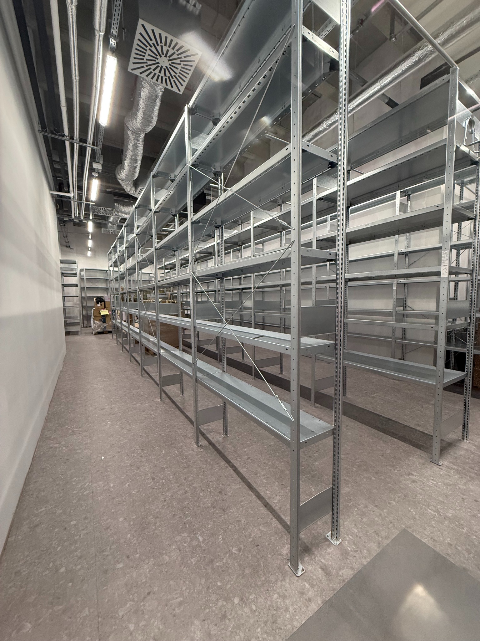 Półki magazynowe z metalu w hali przemysłowej, widok z perspektywy podłogi, oświetlenie sufitowe, osoba w oddali. Czysta przestrzeń gotowa do użytku.