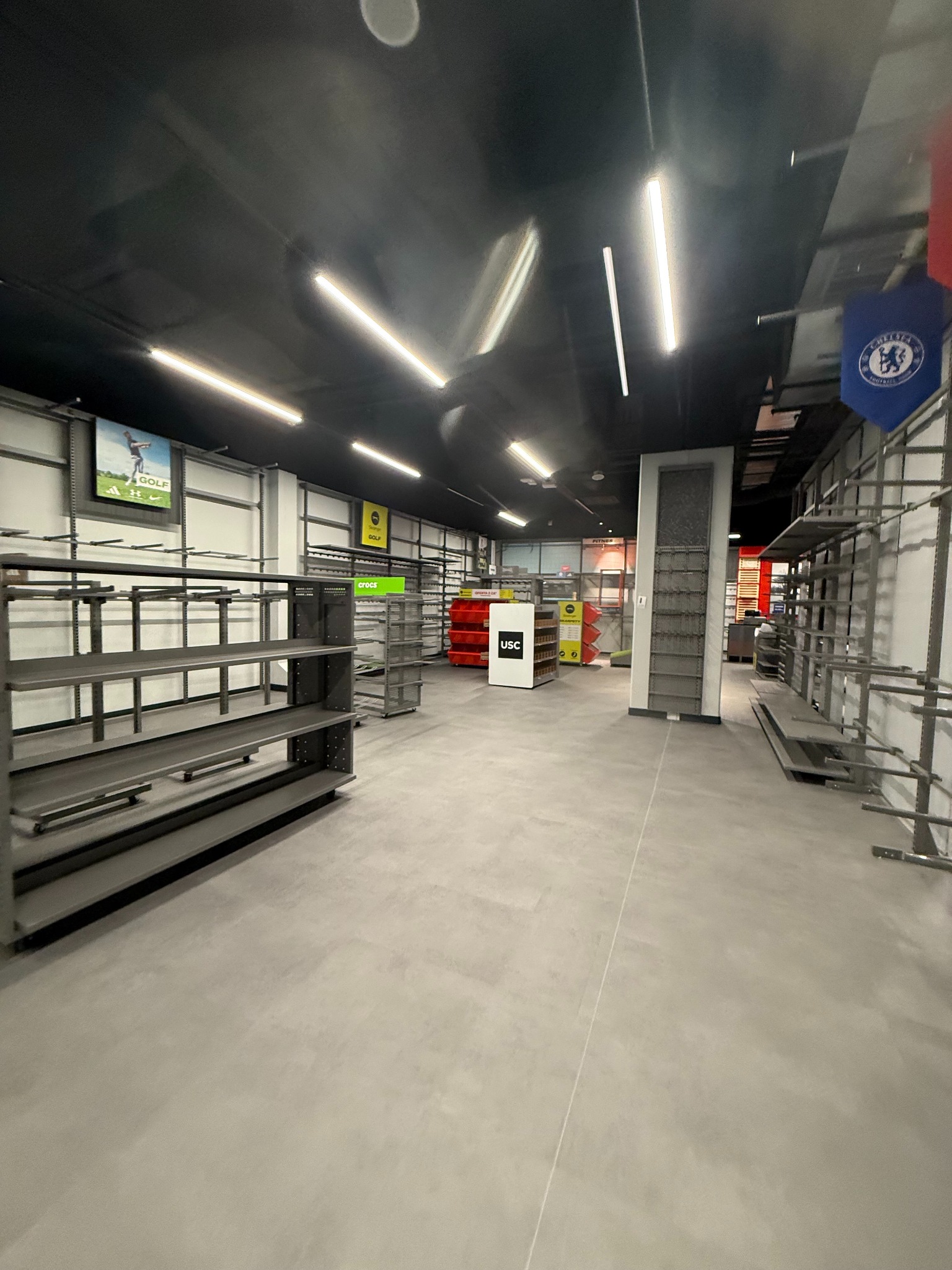 Pusta przestrzeń sklepu z metalowymi regałami i jasną podłogą, oświetlona rzędami lamp. Widoczne logo USC i flaga Chelsea. Przygotowanie do otwarcia lub po remoncie.