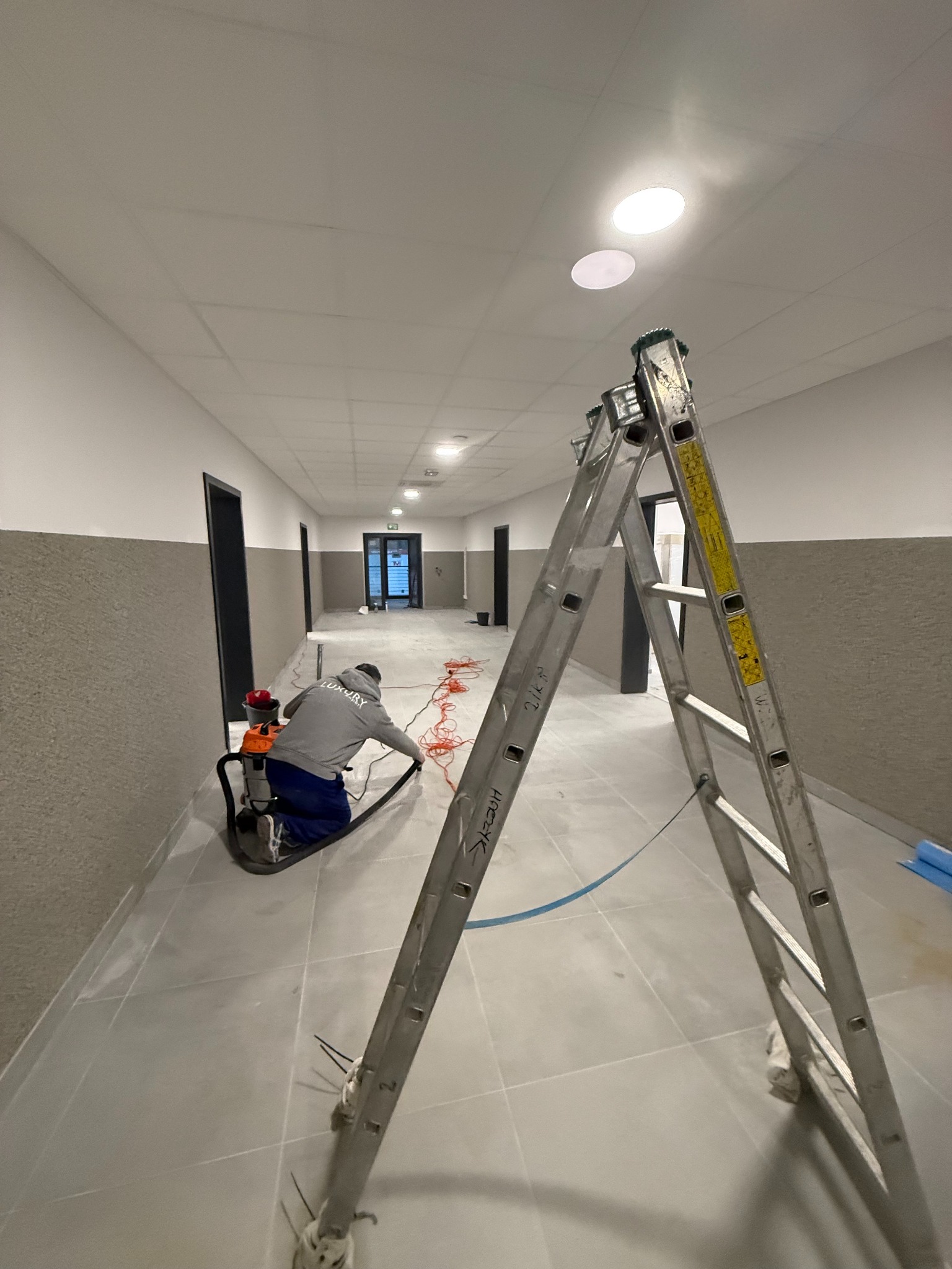 Pracownik w szarym ubraniu czyści podłogę w korytarzu za pomocą odkurzacza przemysłowego. W tle widoczna drabina i drzwi.