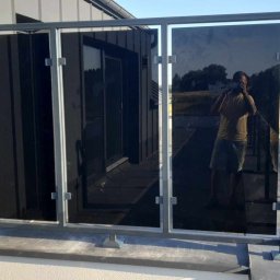 Kamil Aszyk - Szklana balustrada balkonowa z ciemnymi szybami, odbicie fotografa w szybie, widok na pole za balustradą.