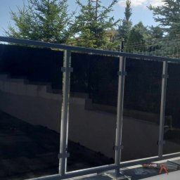 Kamil Aszyk - Nowoczesna balustrada z czarnego szkła i szarej metalowej ramy, zamontowana na niskim murku w ogrodzie, z widocznymi narzędziami montażowymi obok, na tle zieleni i ogrodzenia panelowego.