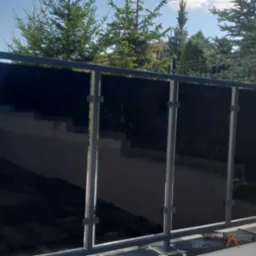 Nowoczesna balustrada z czarnego szkła i szarej metalowej ramy, zamontowana na niskim murku w ogrodzie, z widocznymi narzędziami montażowymi obok, na tle zieleni i ogrodzenia panelowego.