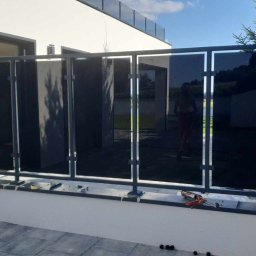 Kamil Aszyk - Nowoczesna balustrada z ciemnego szkła i metalowych ram, zamontowana na tarasie domu jednorodzinnego, widoczne narzędzia montażowe.