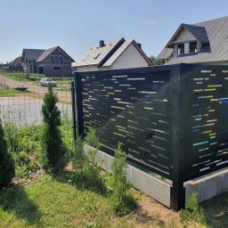 Kamil Aszyk - Nowoczesne ogrodzenie panelowe w kolorze antracytowym z poziomymi, prostokątnymi wycięciami, na tle budynków w stanie surowym otwartym i zieleni.