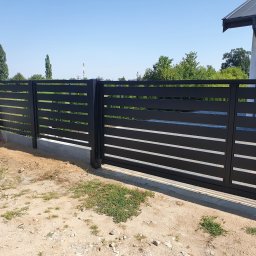 Kamil Aszyk - Nowoczesne, poziome, czarne ogrodzenie panelowe z przesuwną bramą na tle błękitnego nieba i zieleni.