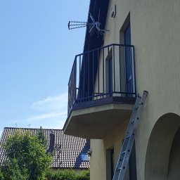 Kamil Aszyk - Czarna balustrada balkonowa z pionowych prętów, widziana z dołu, z anteną na dachu i drabiną opartą o ścianę budynku.