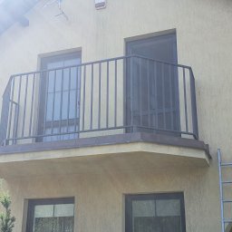 Kamil Aszyk - Metalowa balustrada balkonowa z pionowymi szczeblami, widoczna drabina oparta o ścianę budynku z oknami.