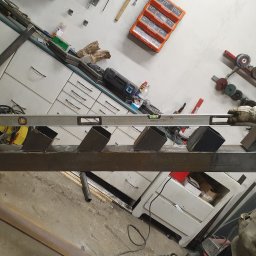 Kamil Aszyk - Spawanie konstrukcji metalowej w warsztacie w Pelplinie: przymiarka metalowych elementów schodów z użyciem poziomicy, widoczne narzędzia i akcesoria warsztatowe w tle.