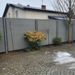 Nowoczesne ogrodzenie panelowe w kolorze szarym z poziomych desek, oddzielające posesję z kostką brukową i roślinnością od domu z ciemną dachówką.