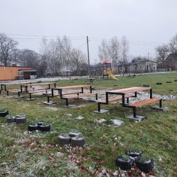 Kamil Aszyk - Zewnętrzna siłownia z drewniano-metalowymi konstrukcjami do ćwiczeń, otoczona trawnikiem z liśćmi i donicami w kształcie pniaków, w tle budynki i plac zabaw, pochmurna pogoda.