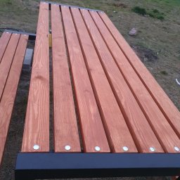 Kamil Aszyk - Nowoczesna ławka parkowa z drewnianym siedziskiem w kolorze brązowym i czarną, metalową konstrukcją, na tle trawnika. Widoczne srebrne śruby mocujące deski.