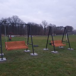Kamil Aszyk - Dwie metalowe huśtawki ogrodowe z drewnianymi siedziskami, zawieszone na łańcuchach, umieszczone na betonowych podstawach na trawiastym terenie.