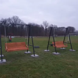 Dwie metalowe huśtawki ogrodowe z drewnianymi siedziskami, zawieszone na łańcuchach, umieszczone na betonowych podstawach na trawiastym terenie.