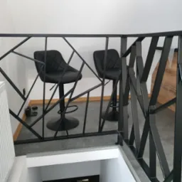 Czarna, metalowa balustrada o geometrycznym wzorze, zabezpieczająca antresolę z widokiem na schody, w tle dwa czarne hokery.