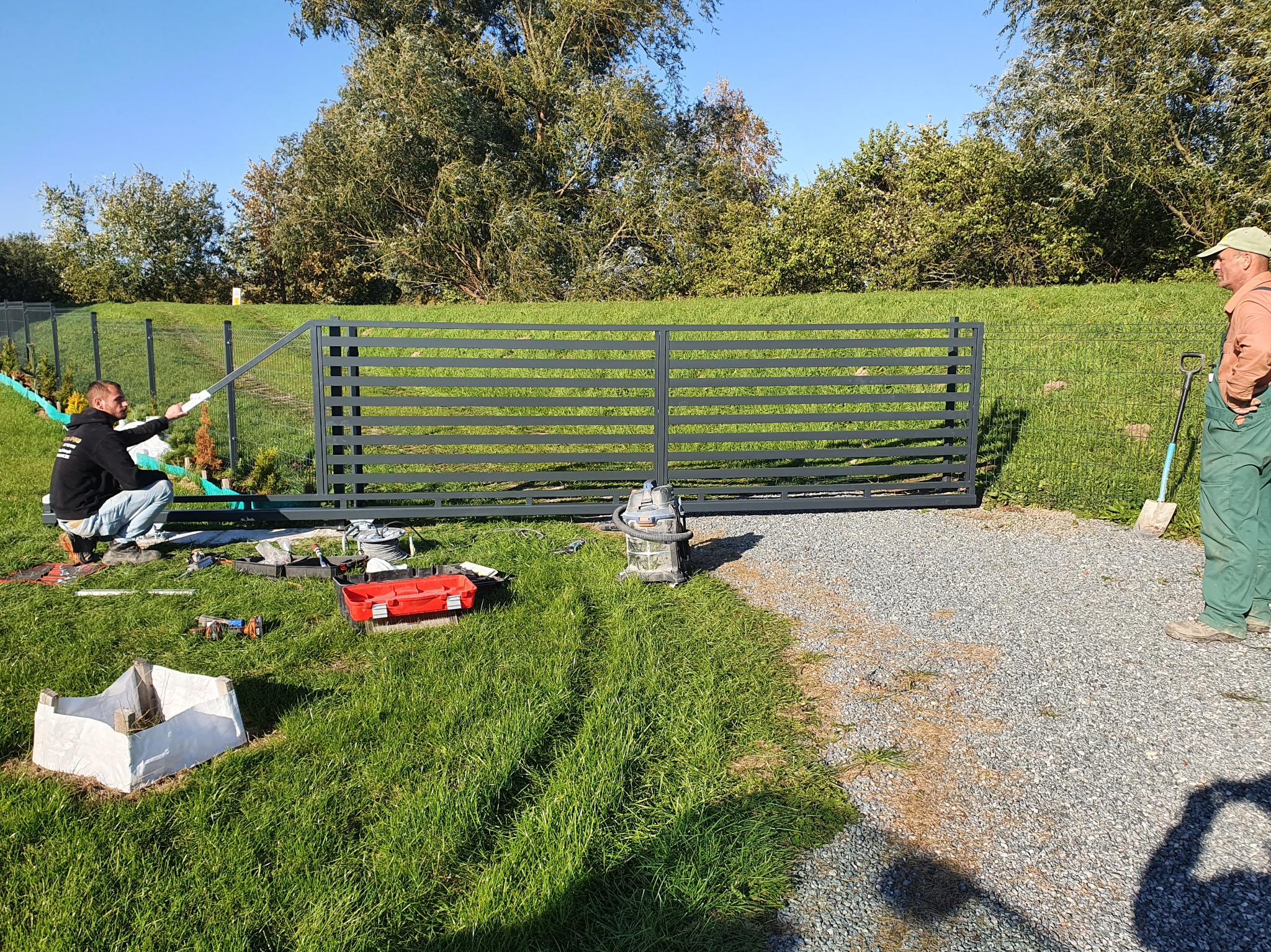 Montaż poziomego ogrodzenia panelowego w ogrodzie, widoczni dwaj mężczyźni podczas pracy, narzędzia rozłożone na trawie