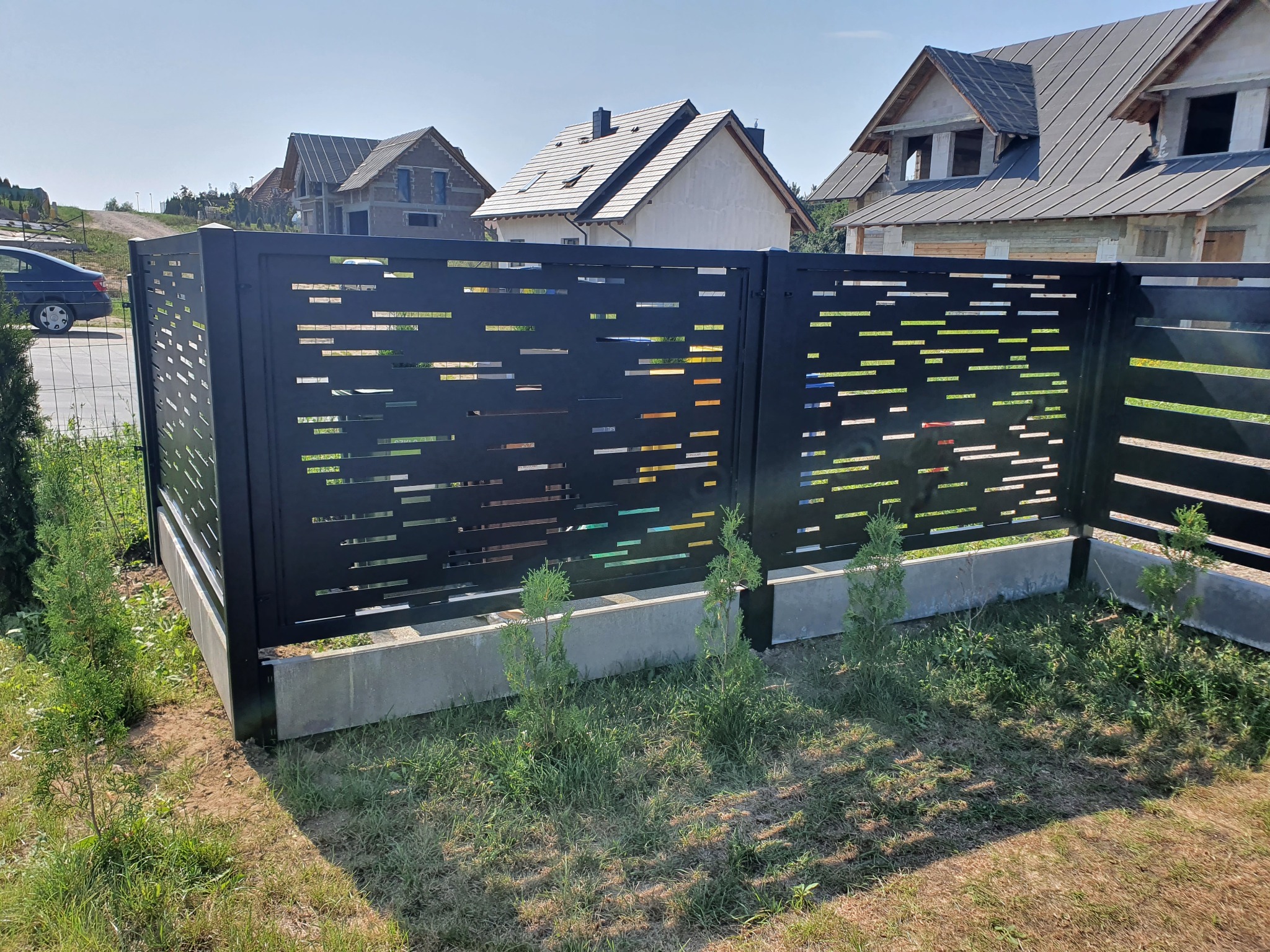 Nowoczesne ogrodzenie panelowe w kolorze czarnym z poziomymi wycięciami, osadzone na betonowej podmurówce, otoczone młodymi tujami i trawą na tle domów w trakcie budowy.