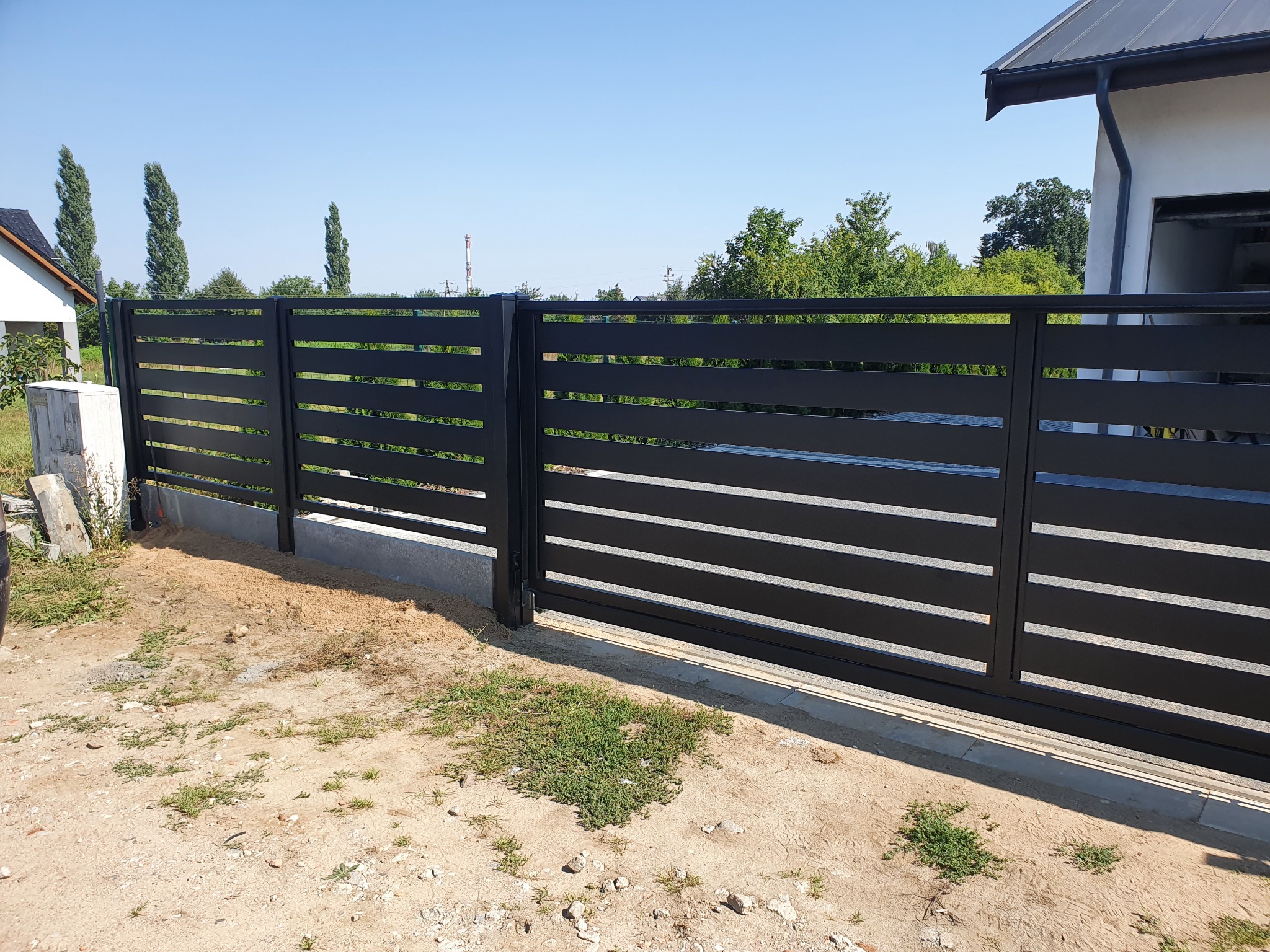 Nowoczesne, poziome, czarne ogrodzenie panelowe z przesuwną bramą na tle błękitnego nieba i zieleni.