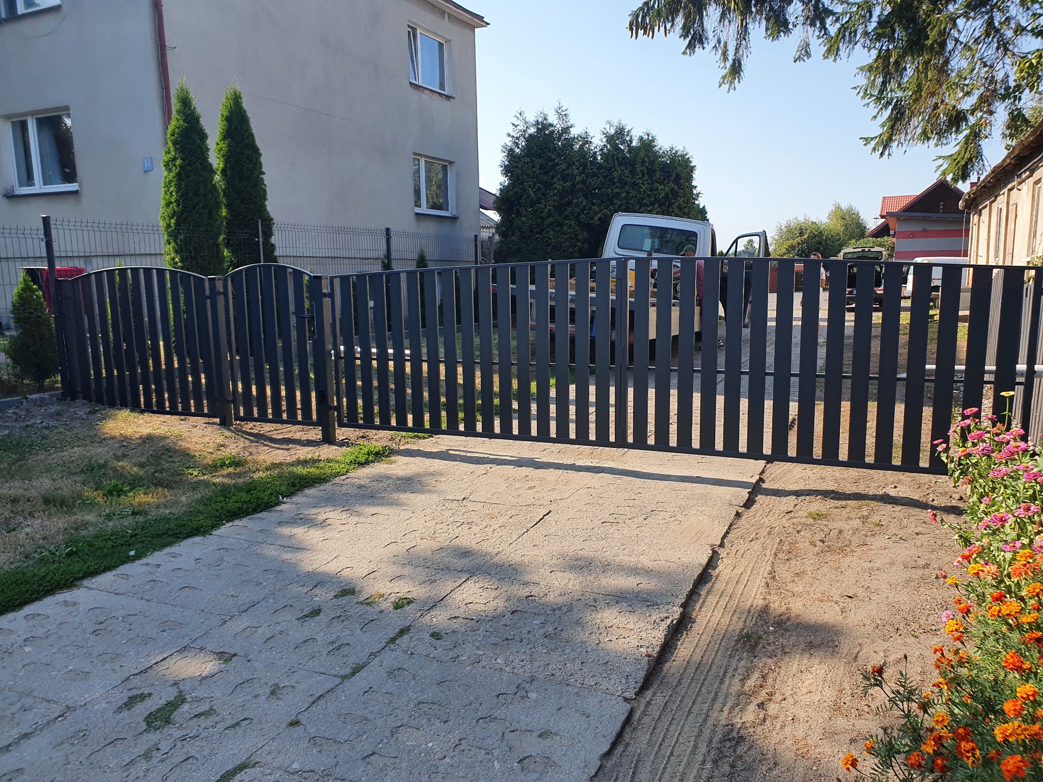 Nowoczesna, grafitowa brama panelowa z furtką na posesji przed domem jednorodzinnym, widoczny fragment chodnika z betonowych płyt, w tle zaparkowany biały samochód dostawczy.