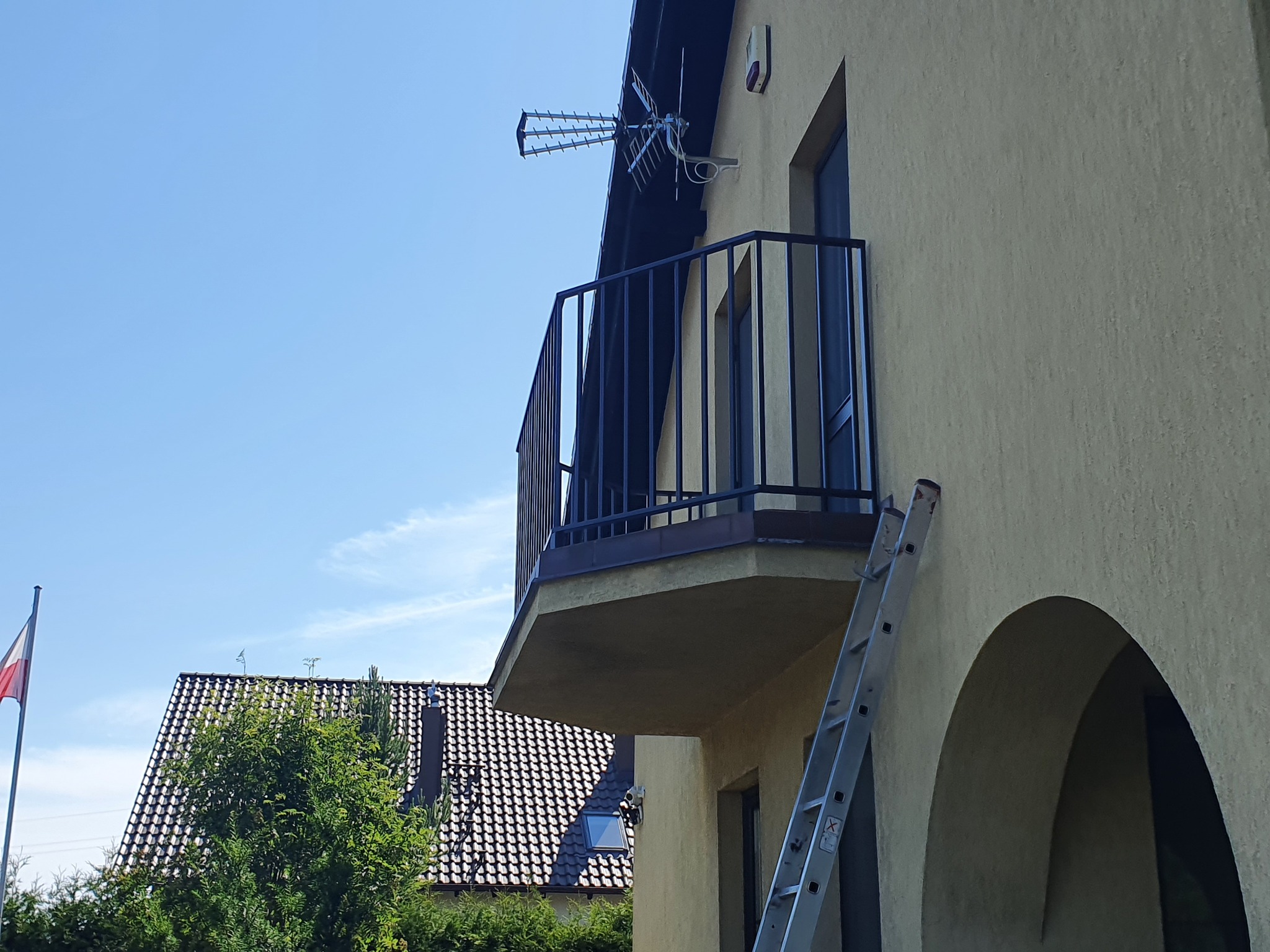 Czarna balustrada balkonowa z pionowych prętów, widziana z dołu, z anteną na dachu i drabiną opartą o ścianę budynku.