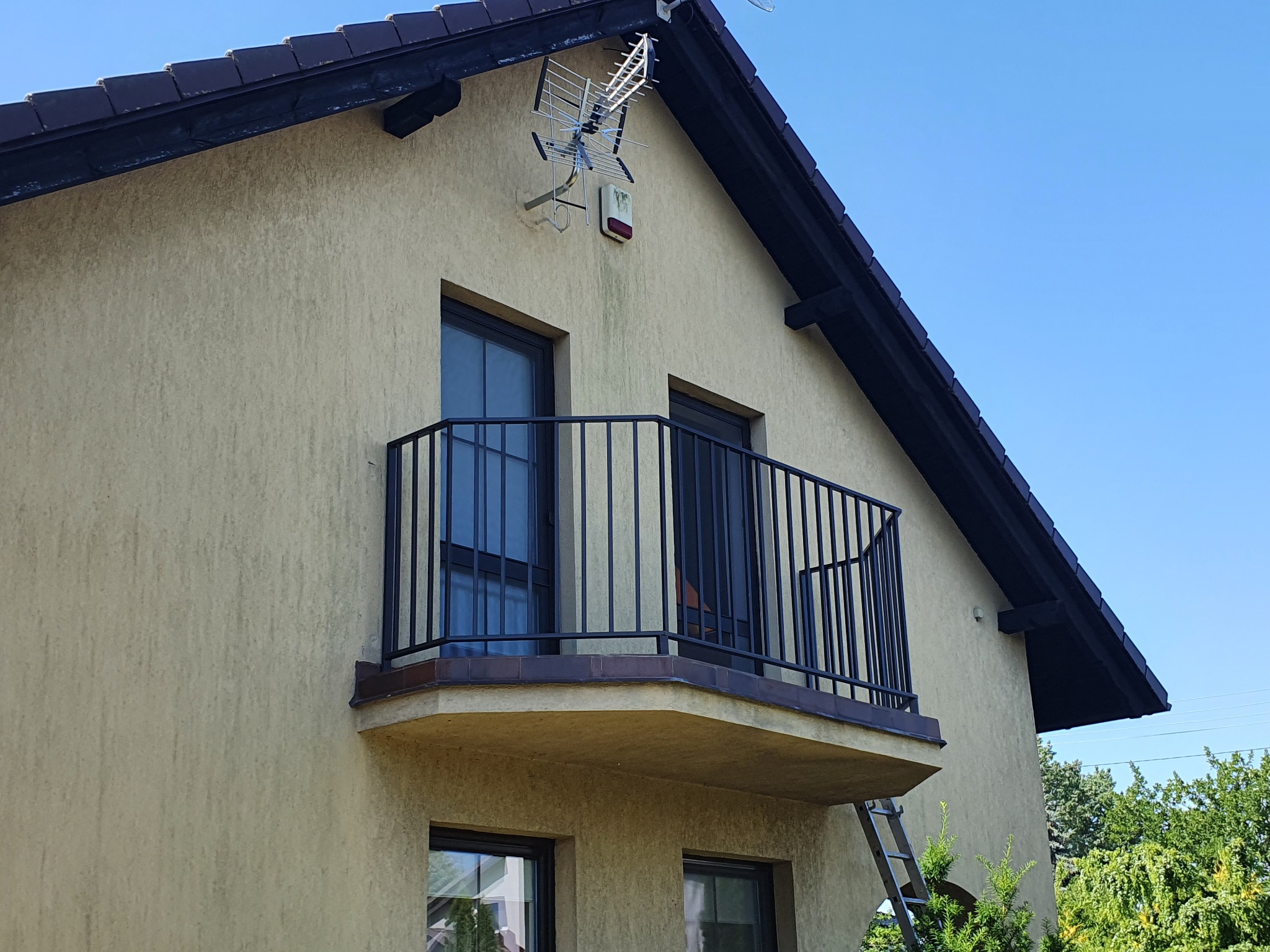Zewnętrzny balkon z czarną metalową balustradą, widoczny z dołu, z drabiną opartą o ścianę budynku.