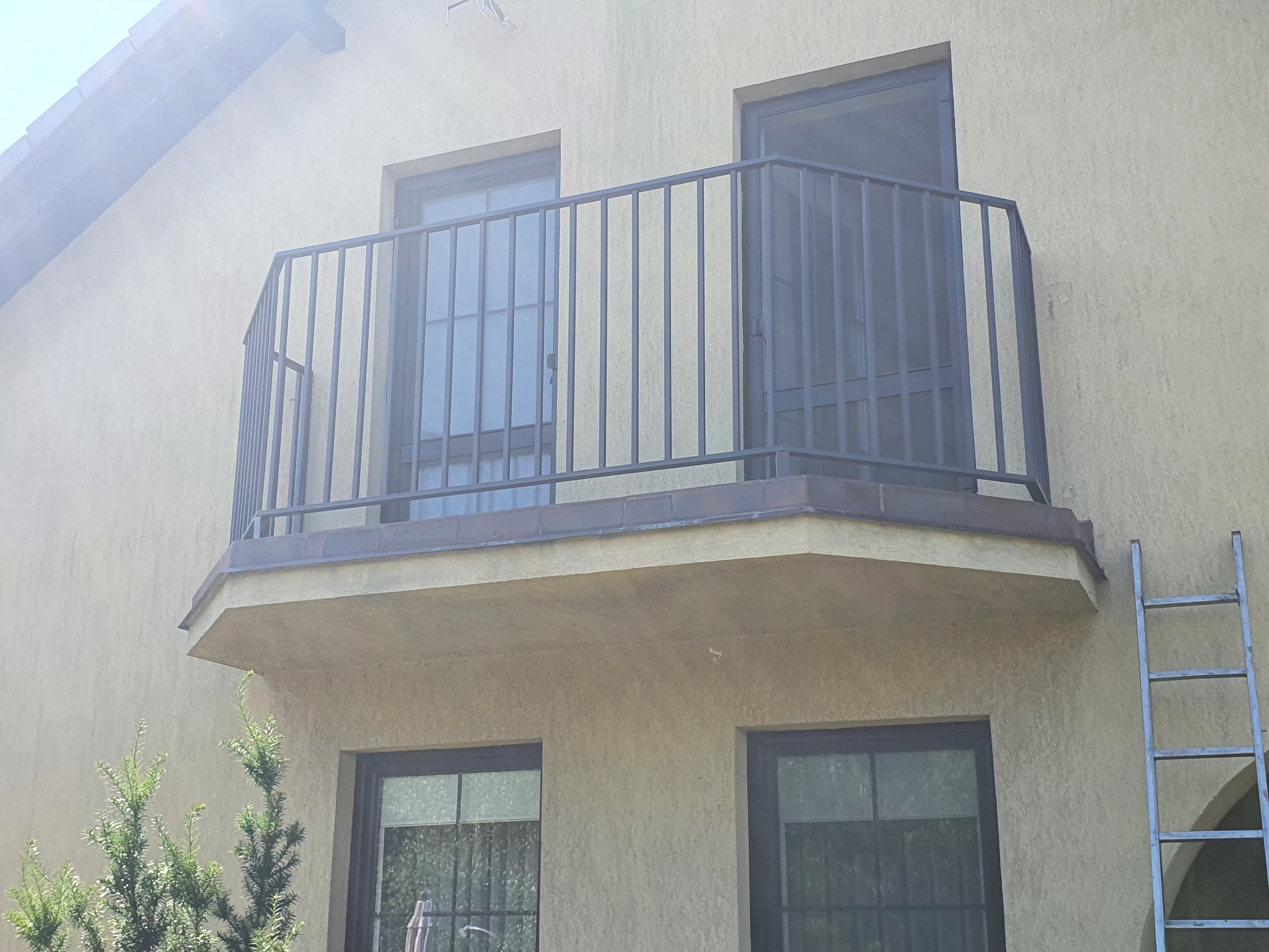 Metalowa balustrada balkonowa z pionowymi prętami, widoczna drabina oparta o ścianę budynku, okna z kratami i małe drzewko iglaste w tle.