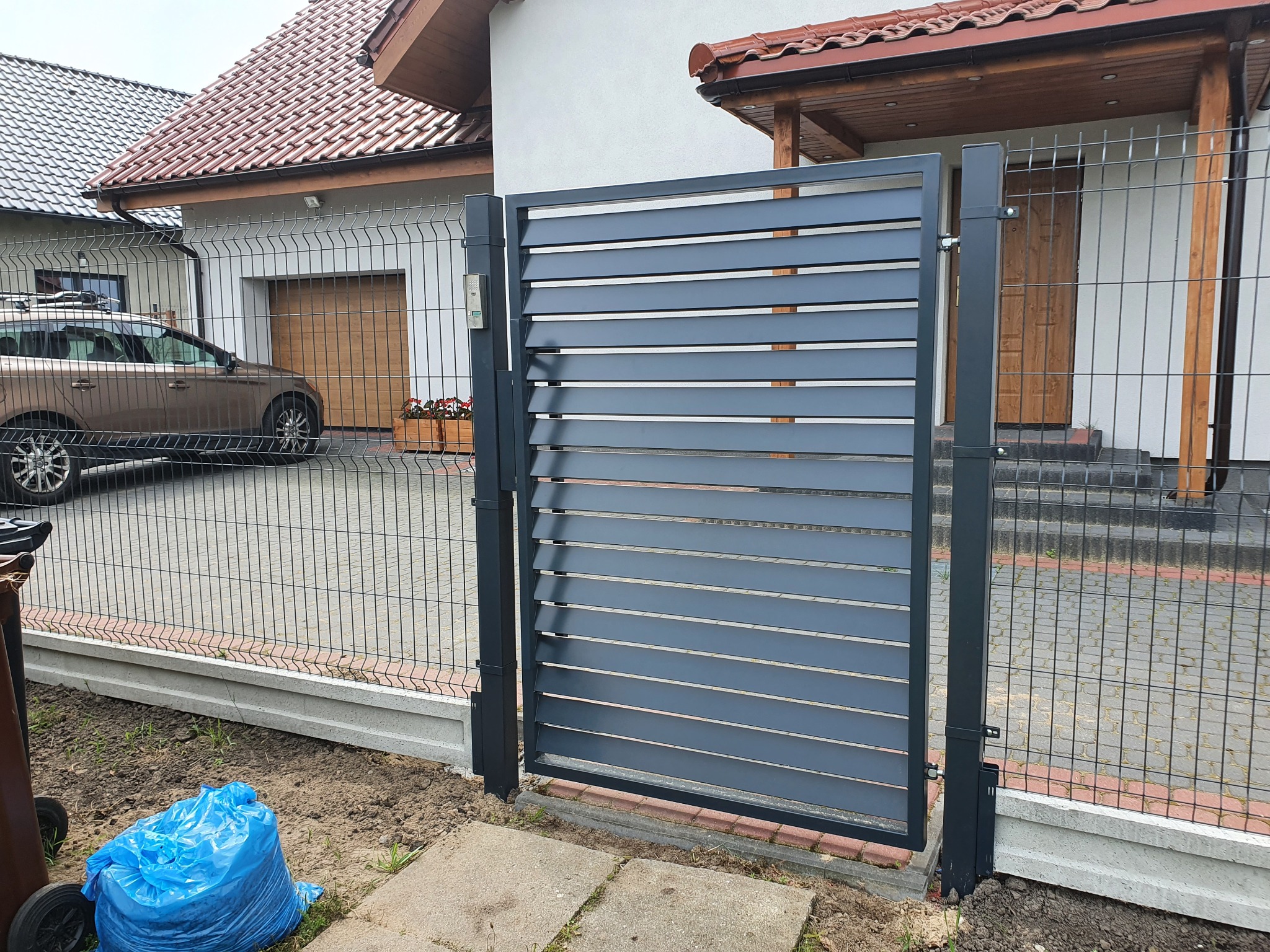 Nowoczesna, grafitowa furtka lamelowa z poziomymi szczeblami, wkomponowana w ogrodzenie panelowe na tle domu z czerwoną dachówką, widoczny fragment posesji i samochód.