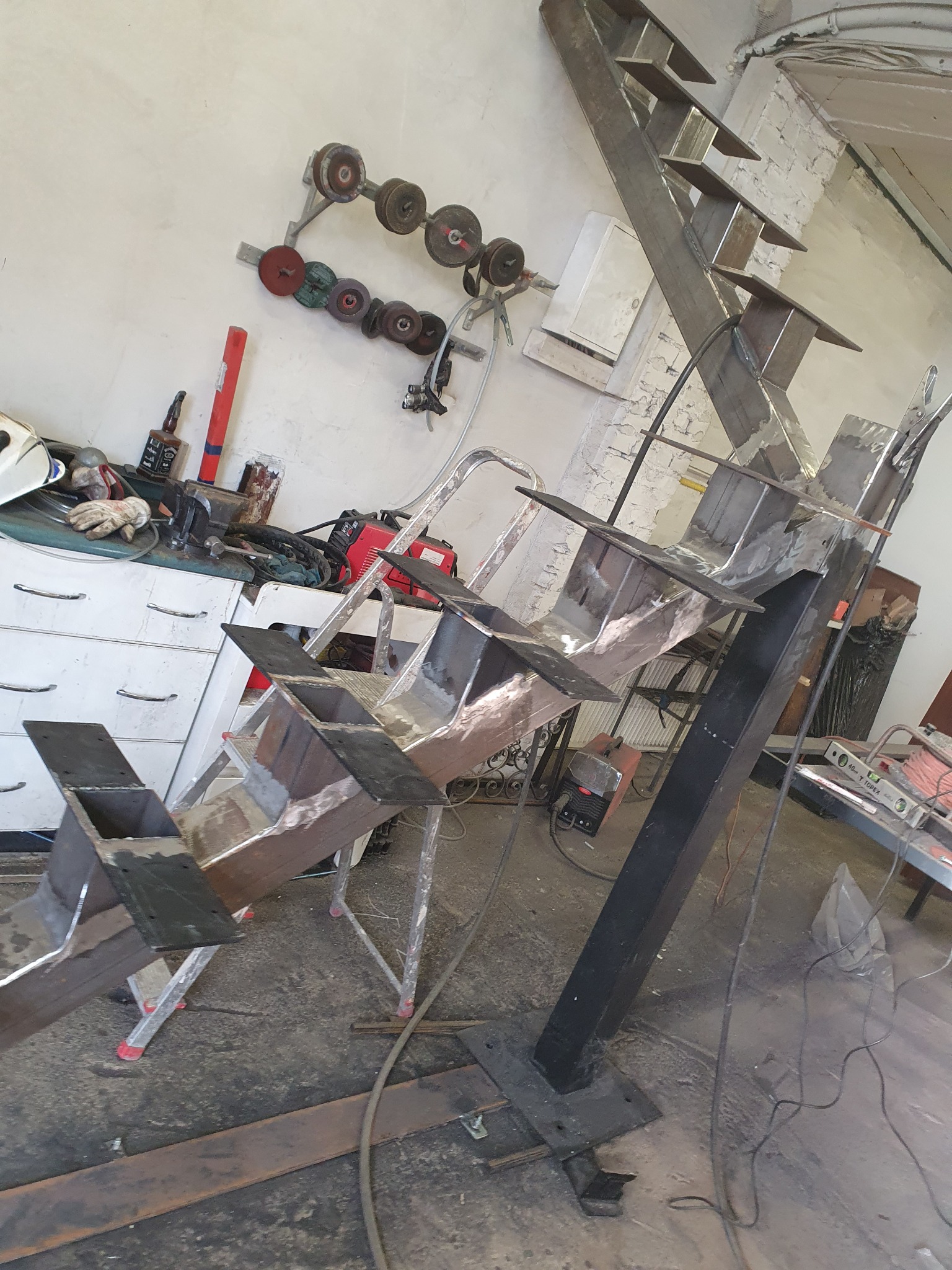 Konstrukcja schodów stalowych w trakcie spawania w warsztacie, widoczne narzędzia i akcesoria spawalnicze, drabina pomocnicza i elementy konstrukcyjne w surowym stanie.