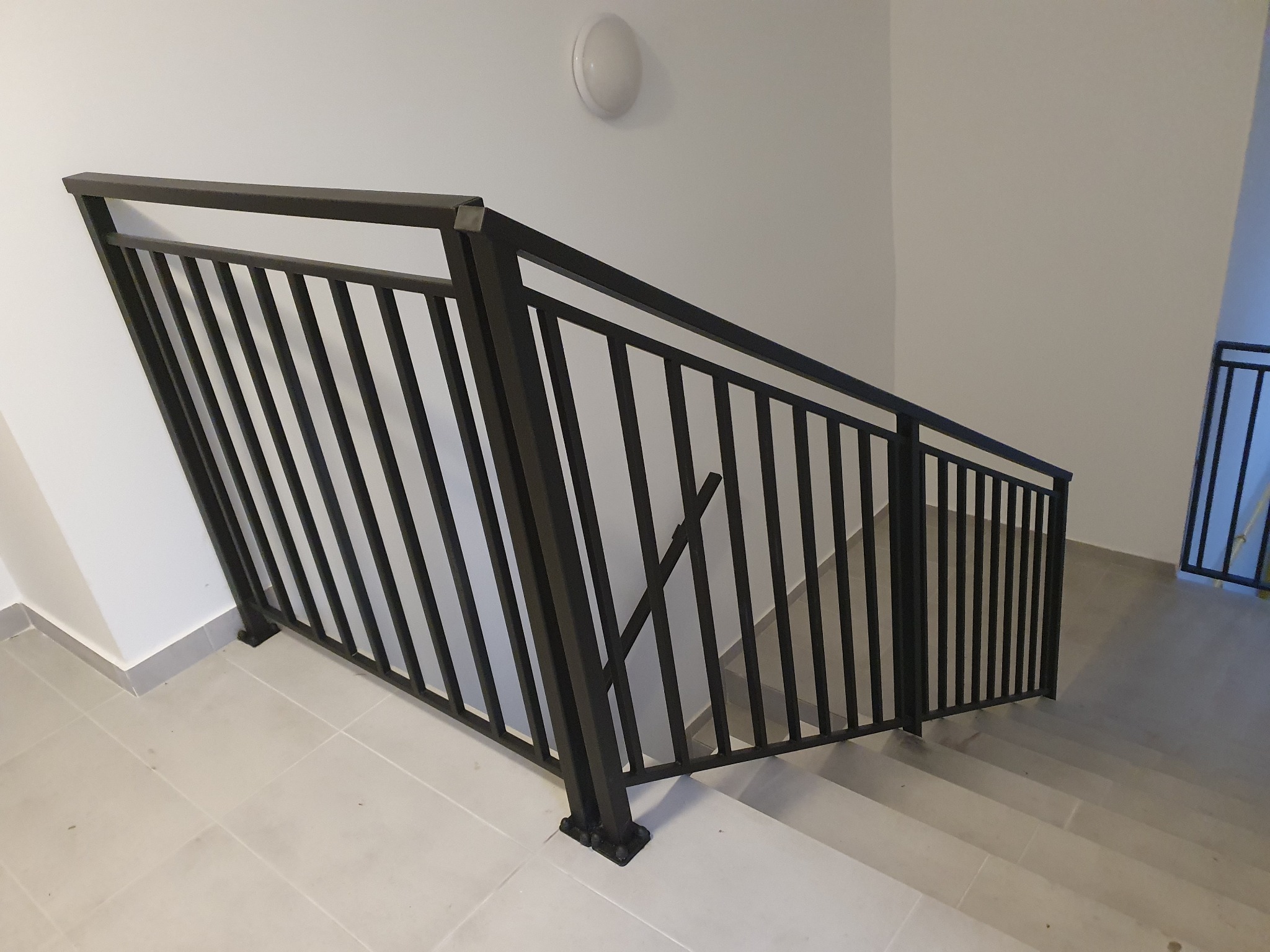 Minimalistyczna, czarna, metalowa balustrada schodowa z pionowymi szczebelkami, zamontowana w jasnym wnętrzu klatki schodowej, widok z dołu na schody pokryte płytkami.