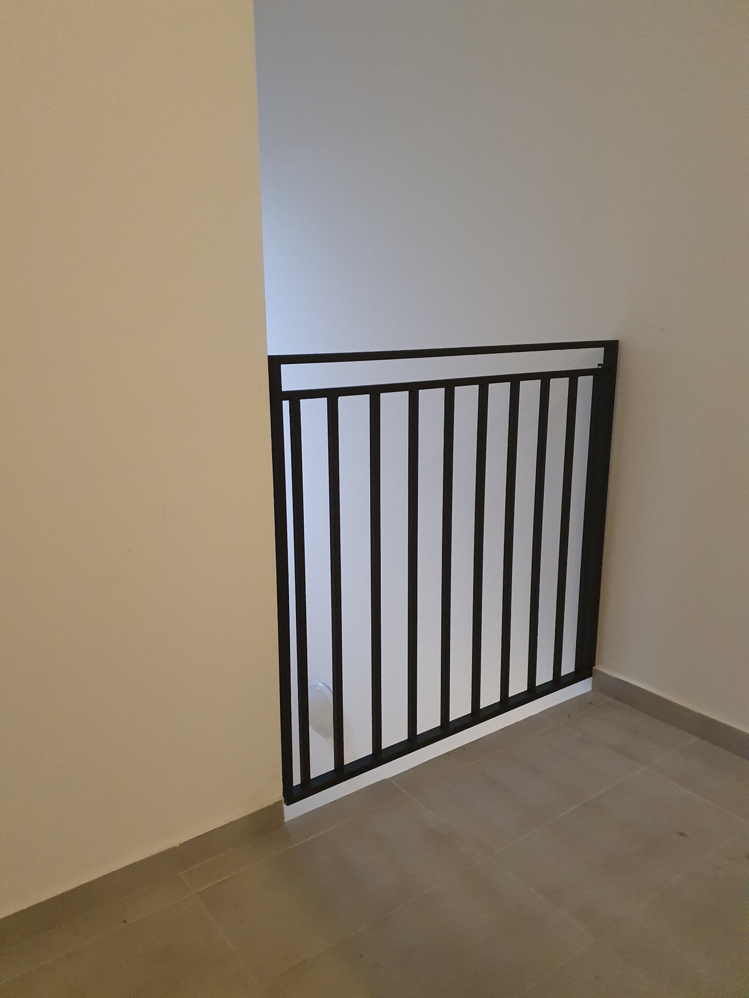Metalowa barierka schodowa z pionowymi szczebelkami, pomalowana na czarno, zamontowana na jasnych płytkach podłogowych wewnątrz budynku.