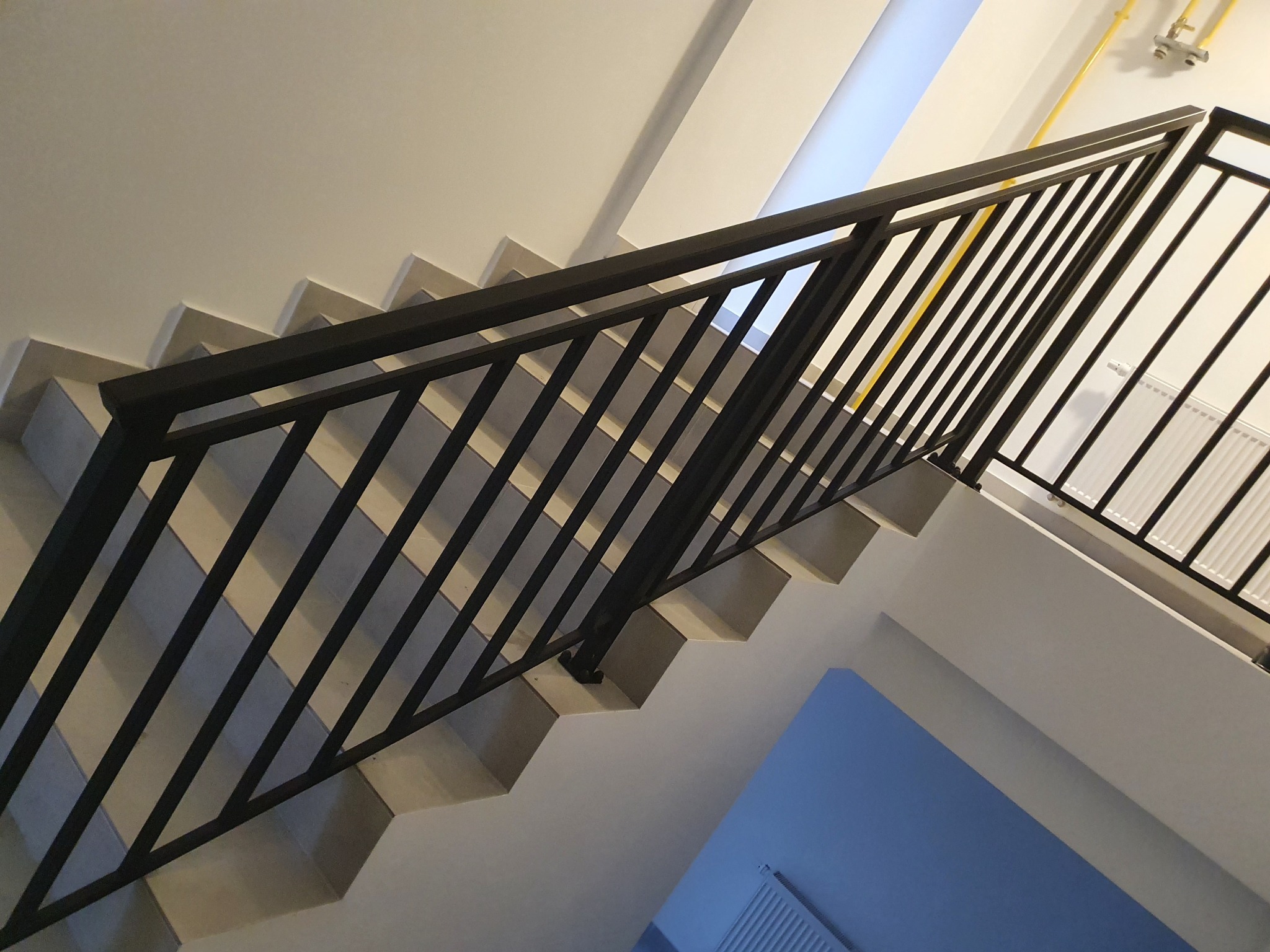 Czarne, metalowe zabezpieczenie schodów z pionowymi prętami, widok z dołu, jasne wnętrze z niebieską ścianą w tle.